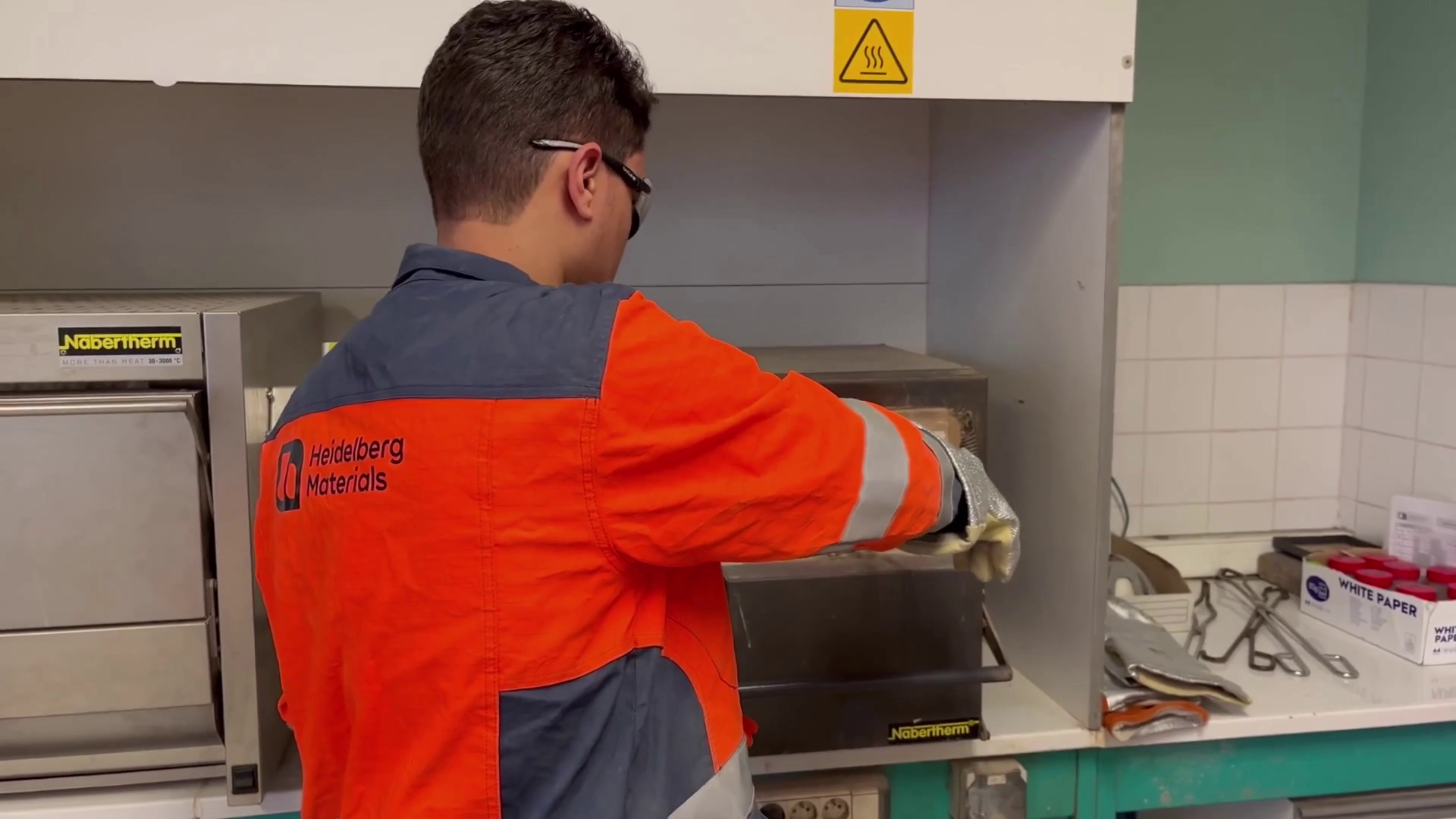 Photo de dos d'un jeune homme brun portant une veste de sécurité orange et bleue avec le logo Heidelberg Materials. Le jeune homme est en train d'effectuer une manipulation. La photo se situe dans un laboratoire.