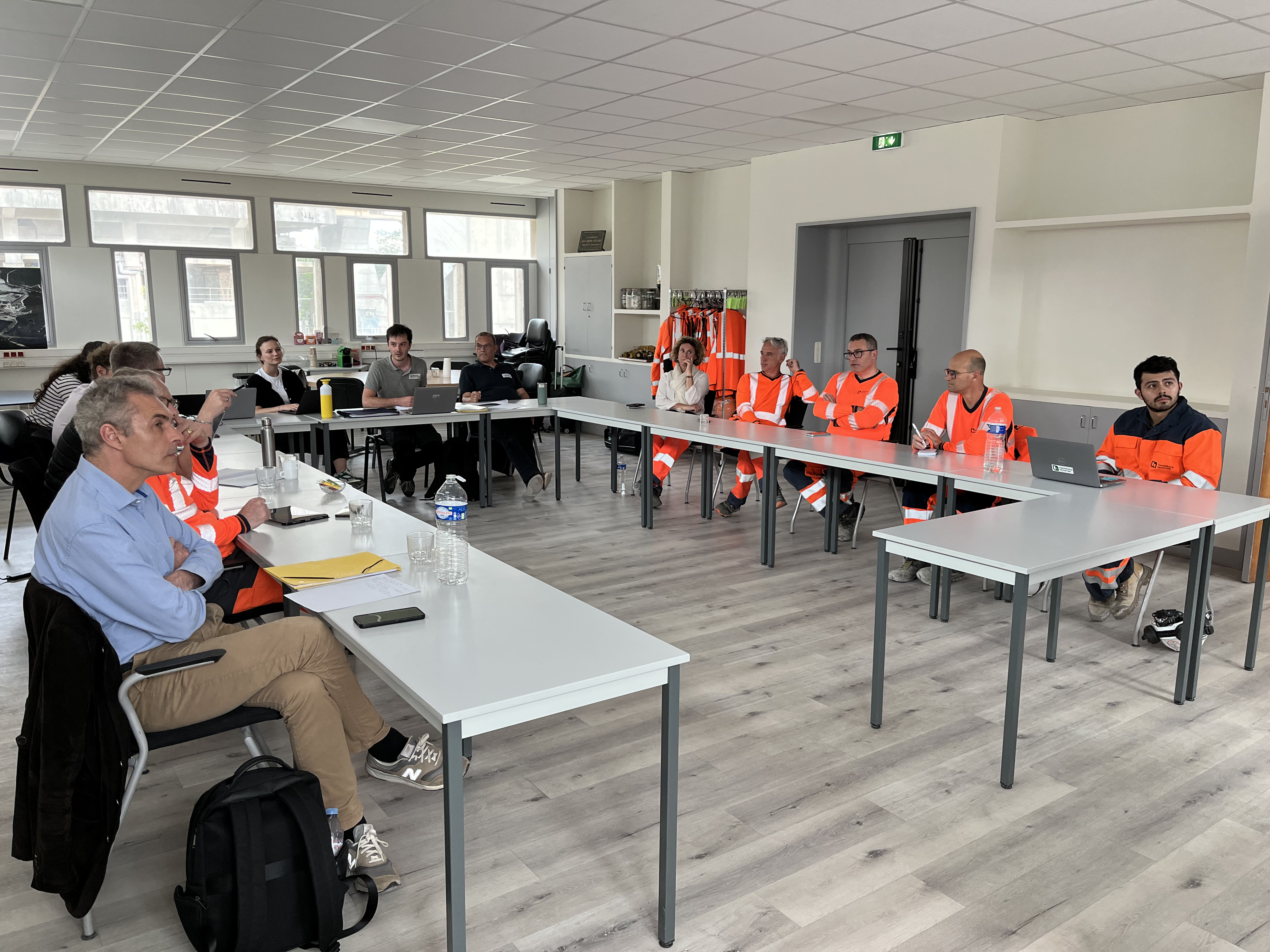 Des personnes assises autour de tables en U dans une salle lumineuse. À gauche, des civils prennent des notes, tandis qu’à droite, des travailleurs en uniformes orange fluorescents assistent à une formation ou une réunion. Arrière-plan avec fenêtres, étagères et porte.