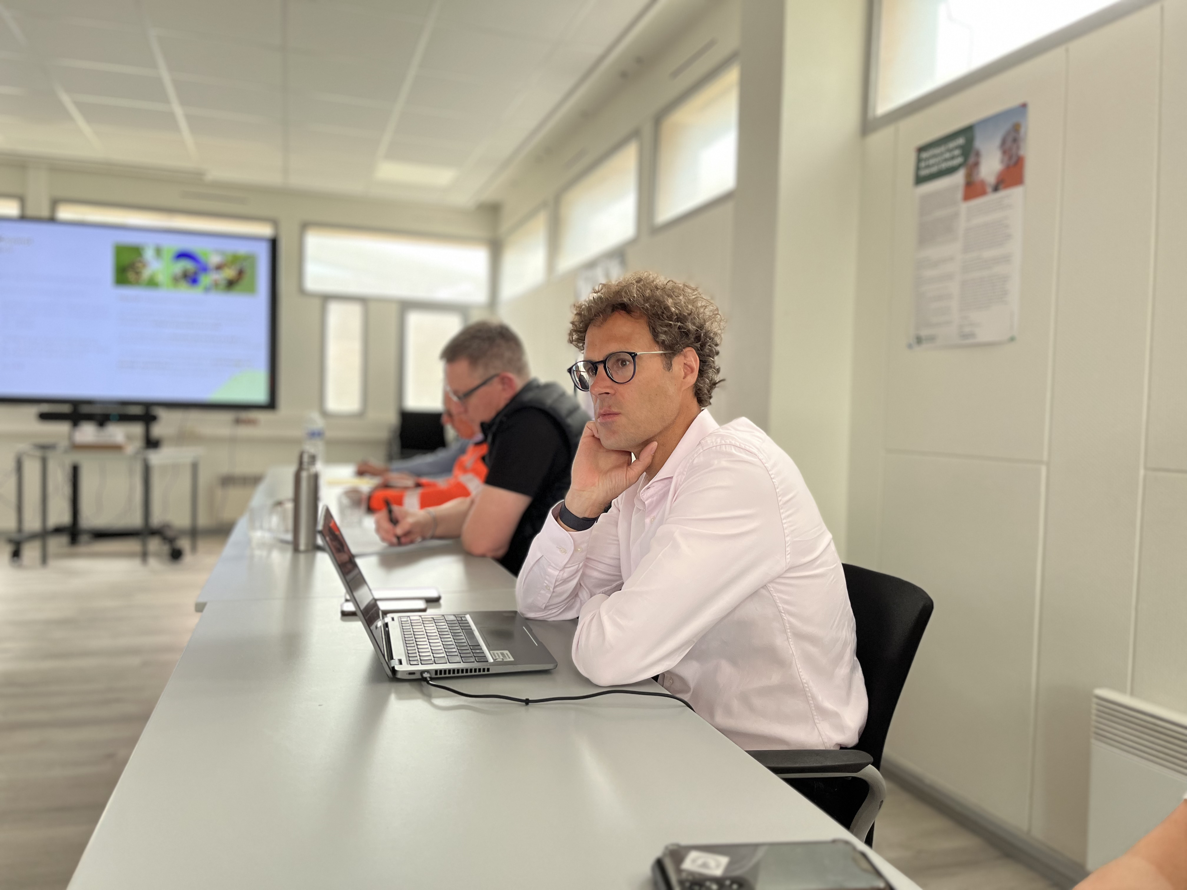 Deux personnes assises à une table de réunion avec des ordinateurs portables, dans une salle lumineuse. Un écran de présentation est visible à l'arrière-plan, affichant des diapositives. Un panneau informatif est accroché au mur à droite.