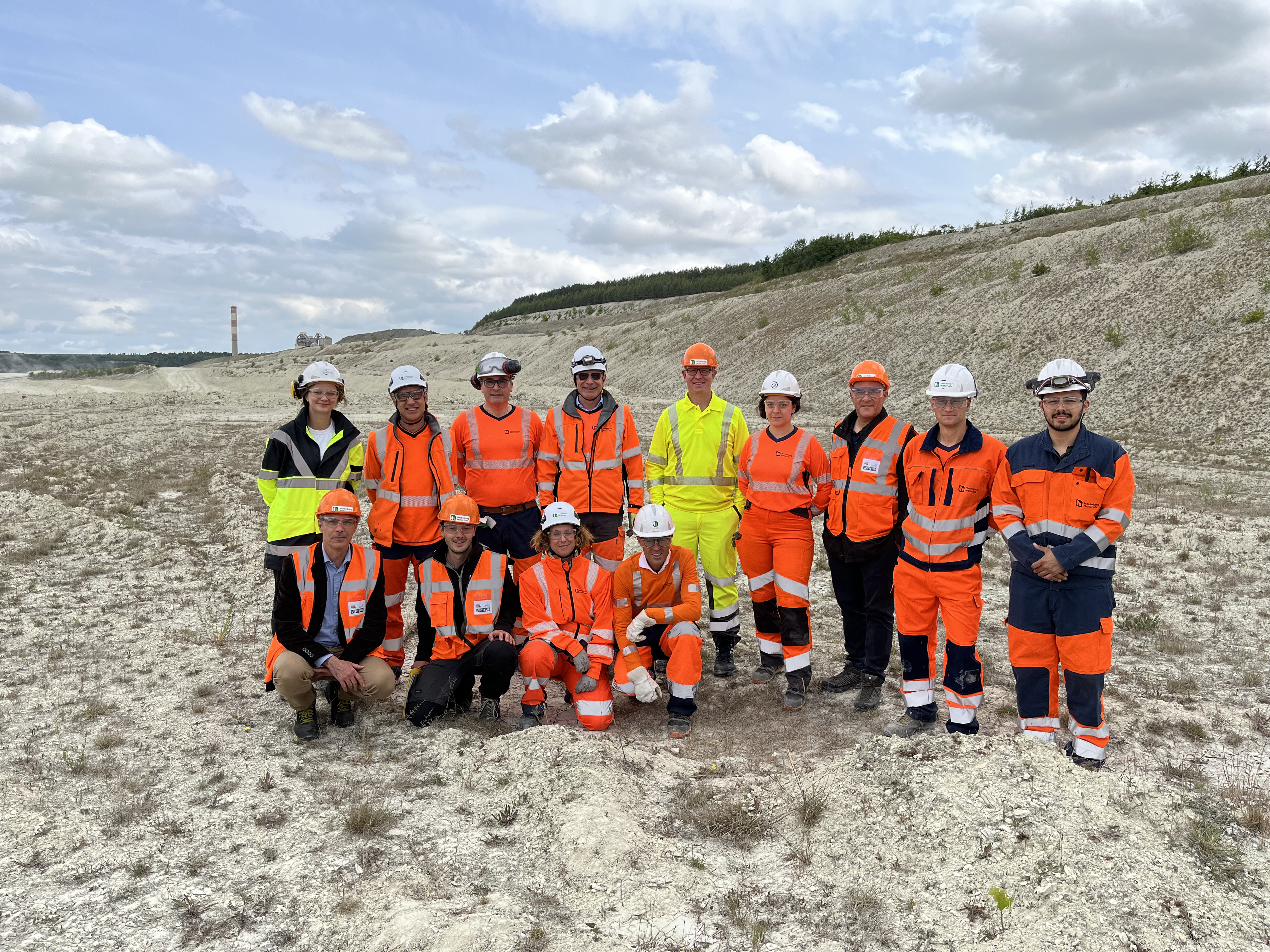 Groupe de 13 personnes en tenue de sécurité orange, jaune et blanche, debout et accroupies sur un site industriel ou une carrière, avec un ciel partiellement nuageux et une cheminée visible à l'horizon sur la gauche.