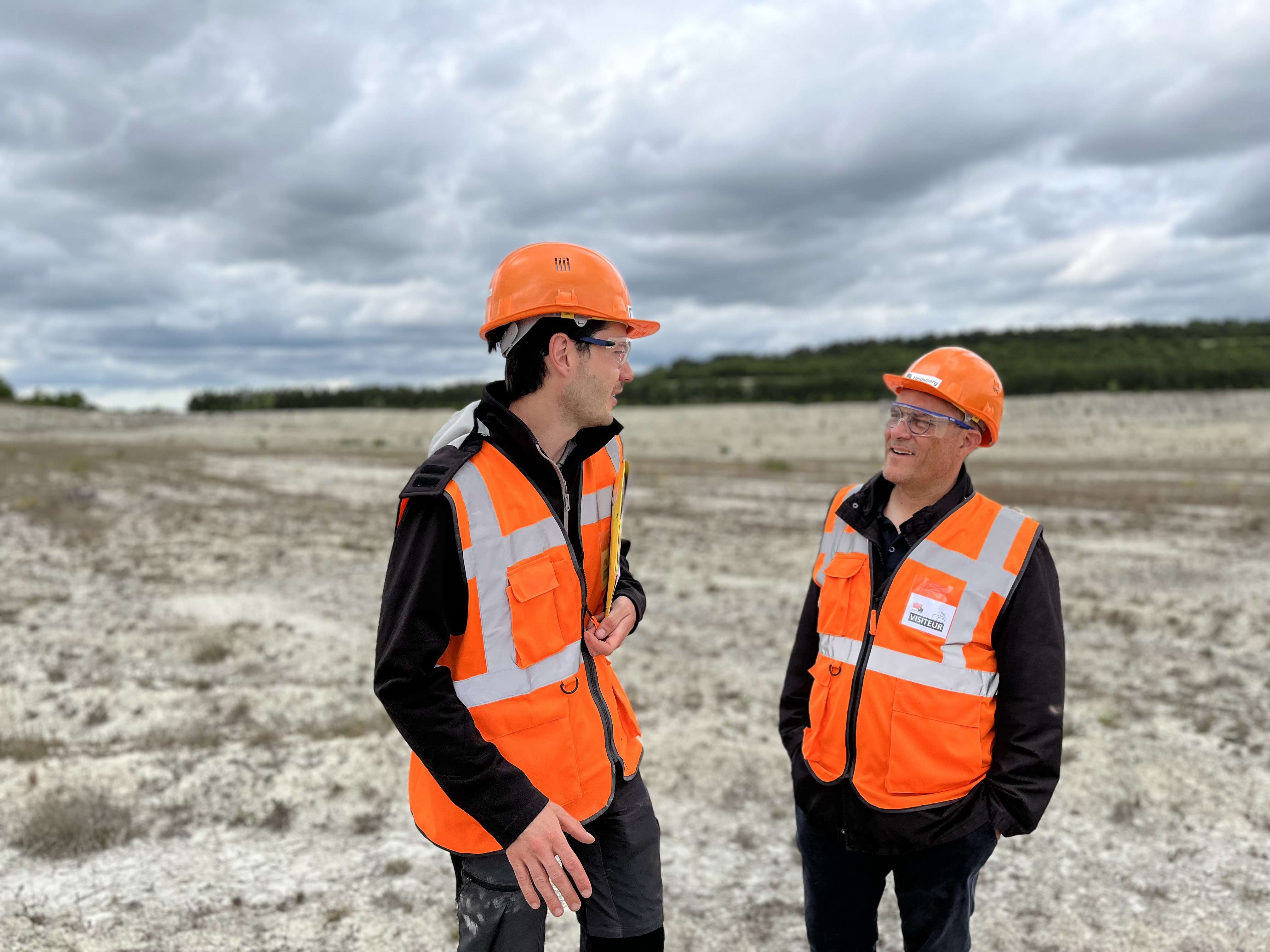 Deux hommes portant des casques et gilets de sécurité orange discutent dans une zone extérieure ouverte avec un terrain rocailleux et un ciel nuageux en arrière-plan.