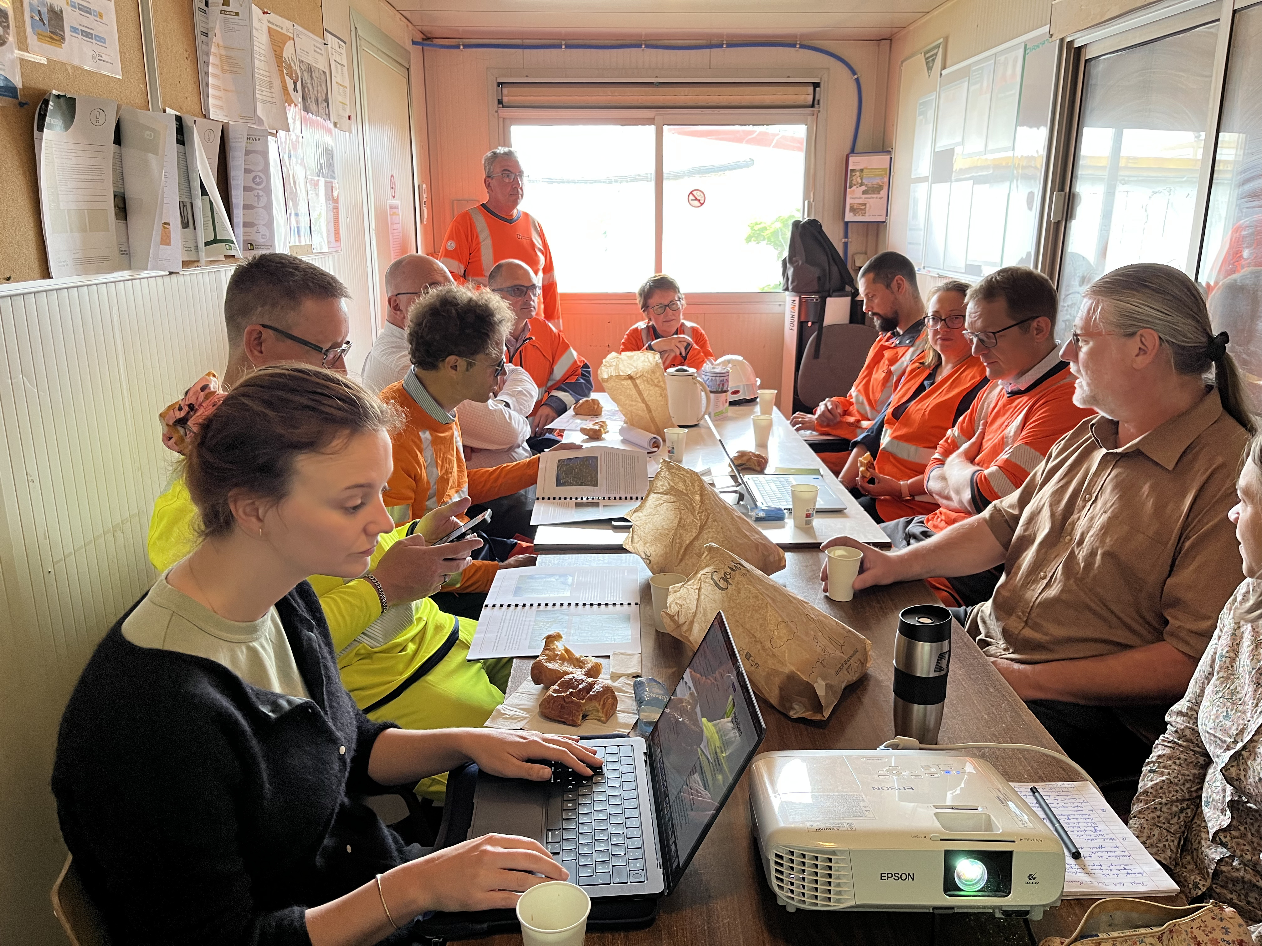 Réunion d'équipe dans une salle de chantier, avec des personnes portant des uniformes orange et d'autres en tenue civile, assises autour d'une table en bois. Sur la table, des ordinateurs portables, des gobelets, des sacs en papier et un projecteur sont visibles. Une grande fenêtre à droite apporte de la lumière naturelle et des documents sont épinglés sur un tableau à gauche. Un homme debout au fond semble s'adresser au groupe.