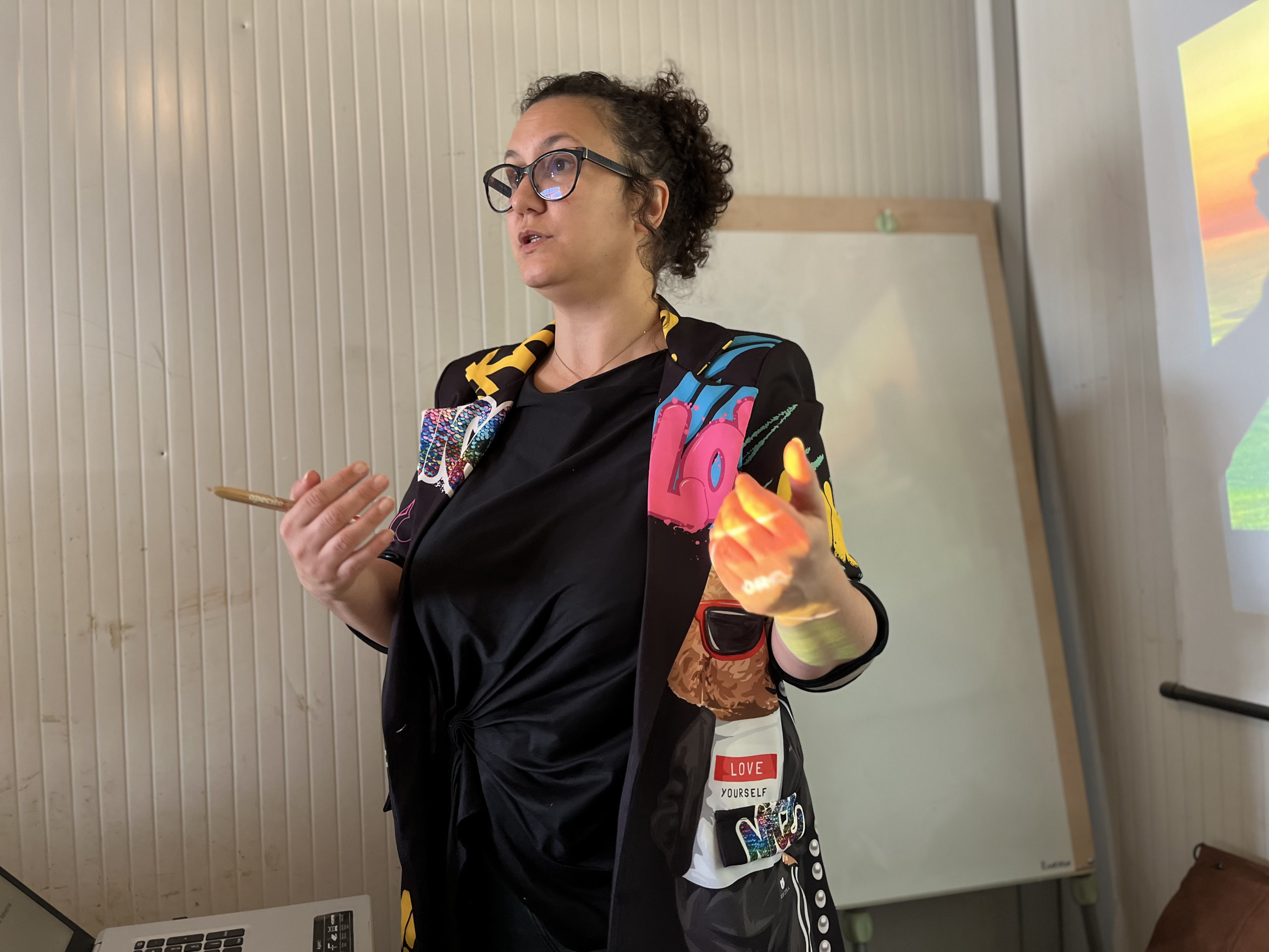 Une personne gesticule en donnant une présentation, tenant un stylo dans une main. En arrière-plan, un tableau blanc et une projection colorée sur un mur à droite. Le cadre induit un contexte éducatif ou professionnel.