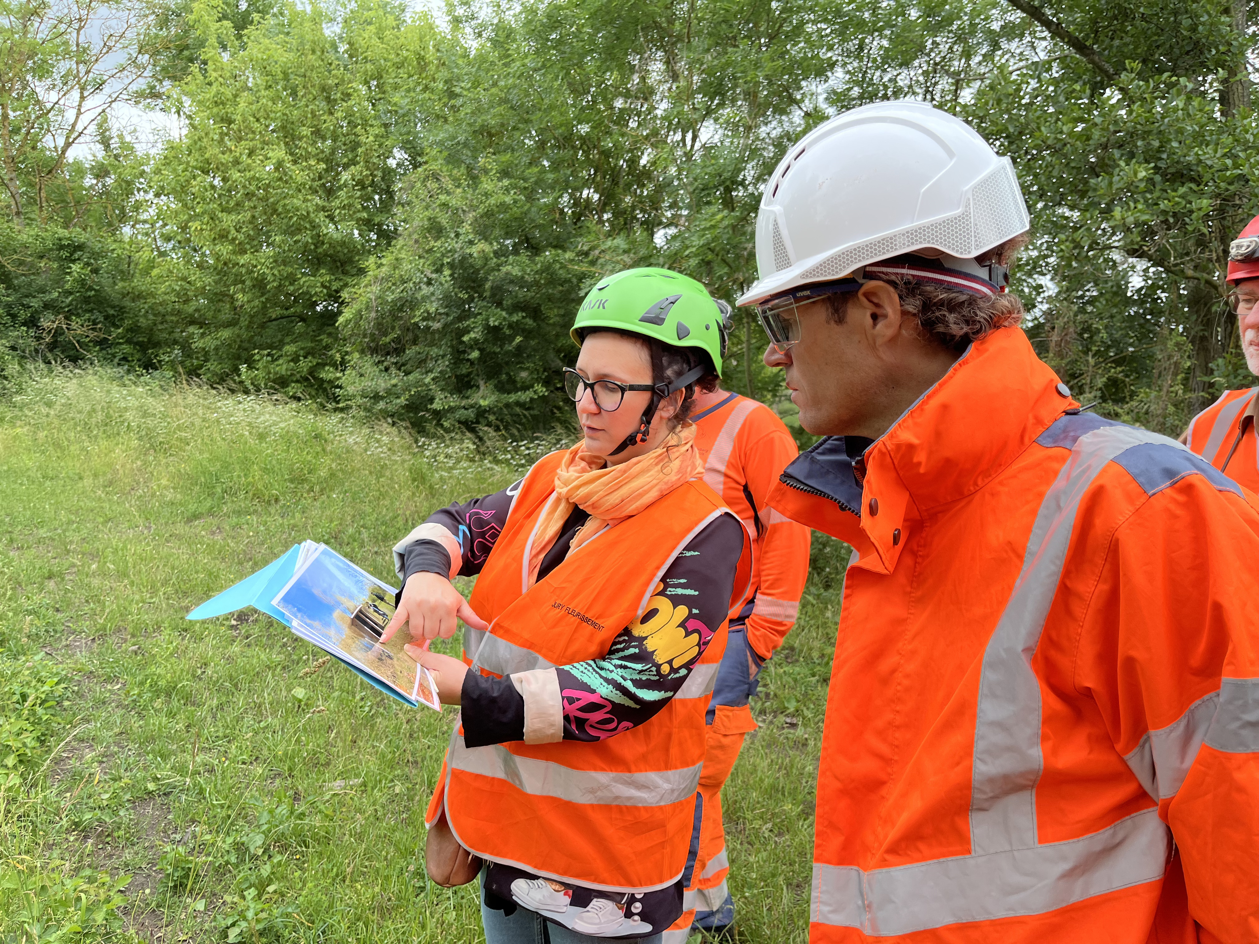 Des travailleurs en gilets orange et casques de sécurité discutent en plein air près d'une zone herbeuse; l'un d'eux montre une carte ou un plan, probablement pour expliquer un projet ou un site.