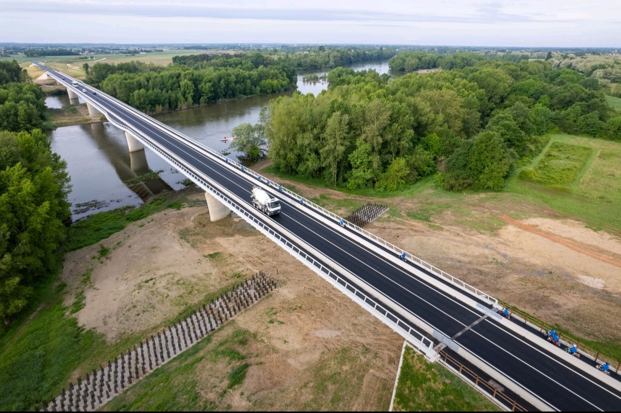 Vue aérienne d'un pont moderne à deux voies traversant une rivière entourée de forêts verdoyantes et de prairies. Une camionnette blanche se déplace au centre du pont, et des barrières de sécurité longent les côtés. Les environs montrent un mélange de nature et de terrain dégagé.
