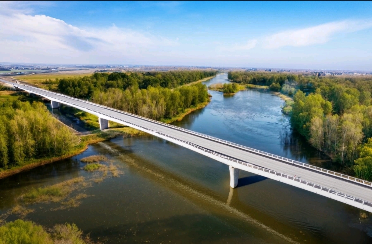 Vue aérienne d'un long pont moderne traversant une rivière entourée de forêts verdoyantes. Le pont est orienté en diagonale, reliant les rives gauche et droite. Le ciel est bleu avec quelques nuages légers en arrière-plan.