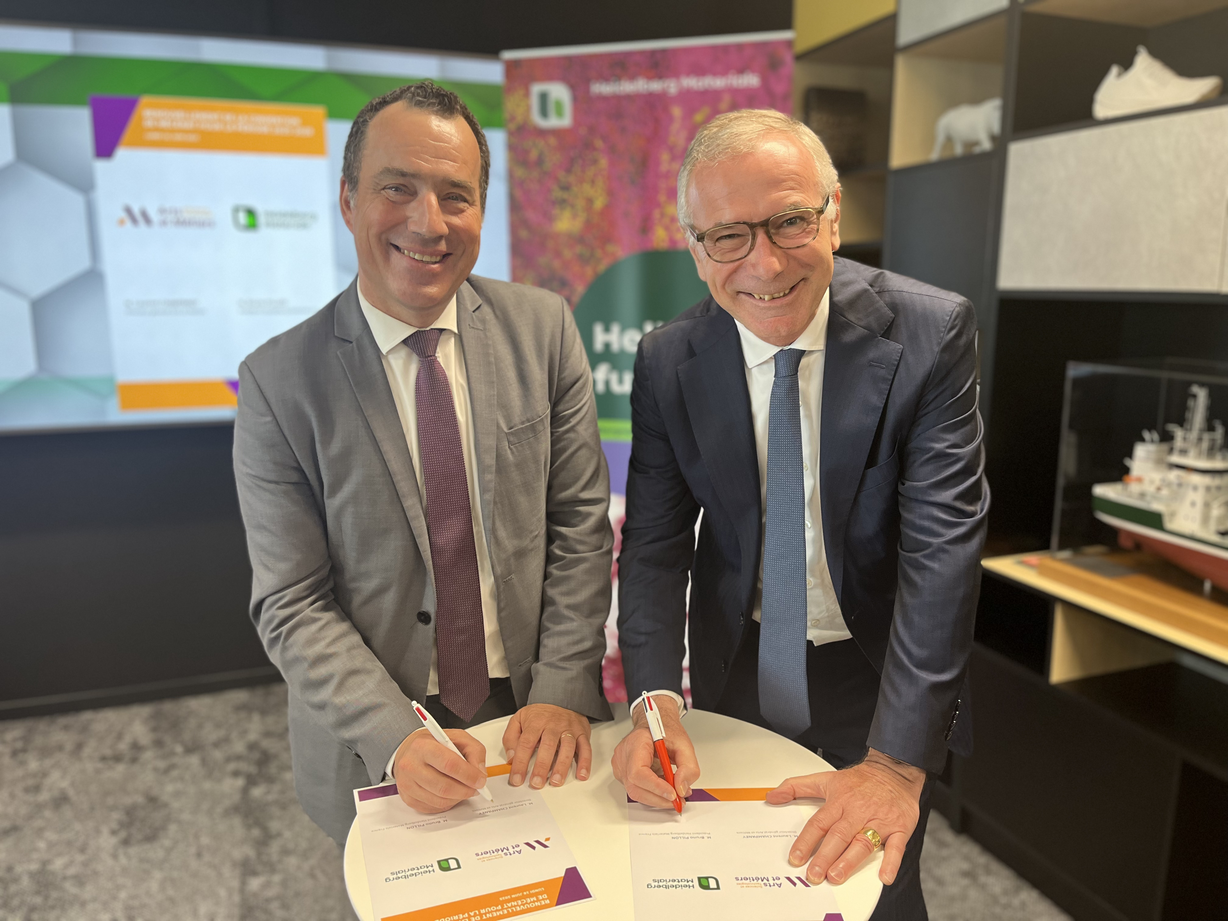 Deux hommes en costumes signent des documents sur une petite table ronde blanche. En arrière-plan, un écran affiche des logos et le titre "Partenariat" avec des éléments graphiques colorés. Sur la droite, une maquette de bateau est exposée sur une étagère.