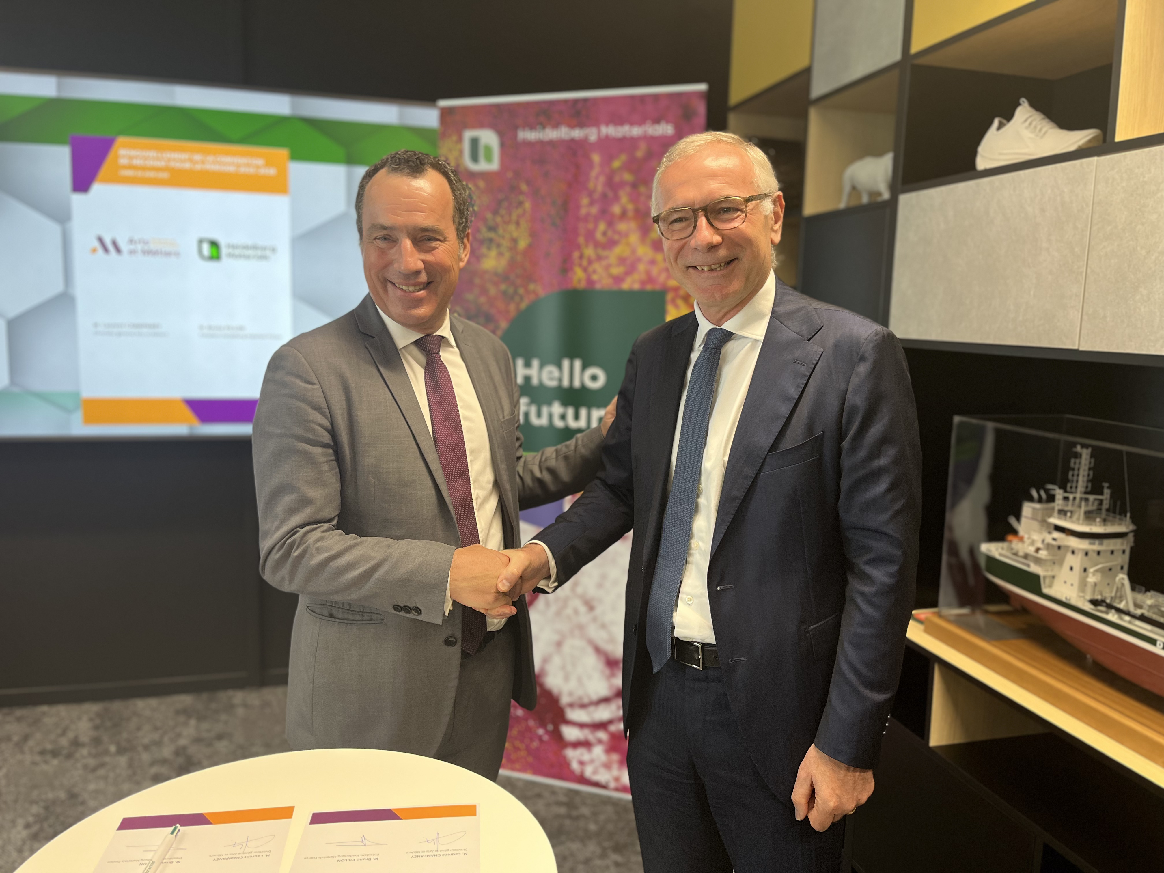 Deux hommes en costume se serrent la main devant une table circulaire blanche avec des documents posés dessus. En arrière-plan, un écran affiche des logos et des informations textuelles, ainsi qu'une bannière multicolore avec le texte "Hello future!". À droite, une étagère contient une maquette de bateau. L'ambiance semble être celle d'une salle de réunion professionnelle.