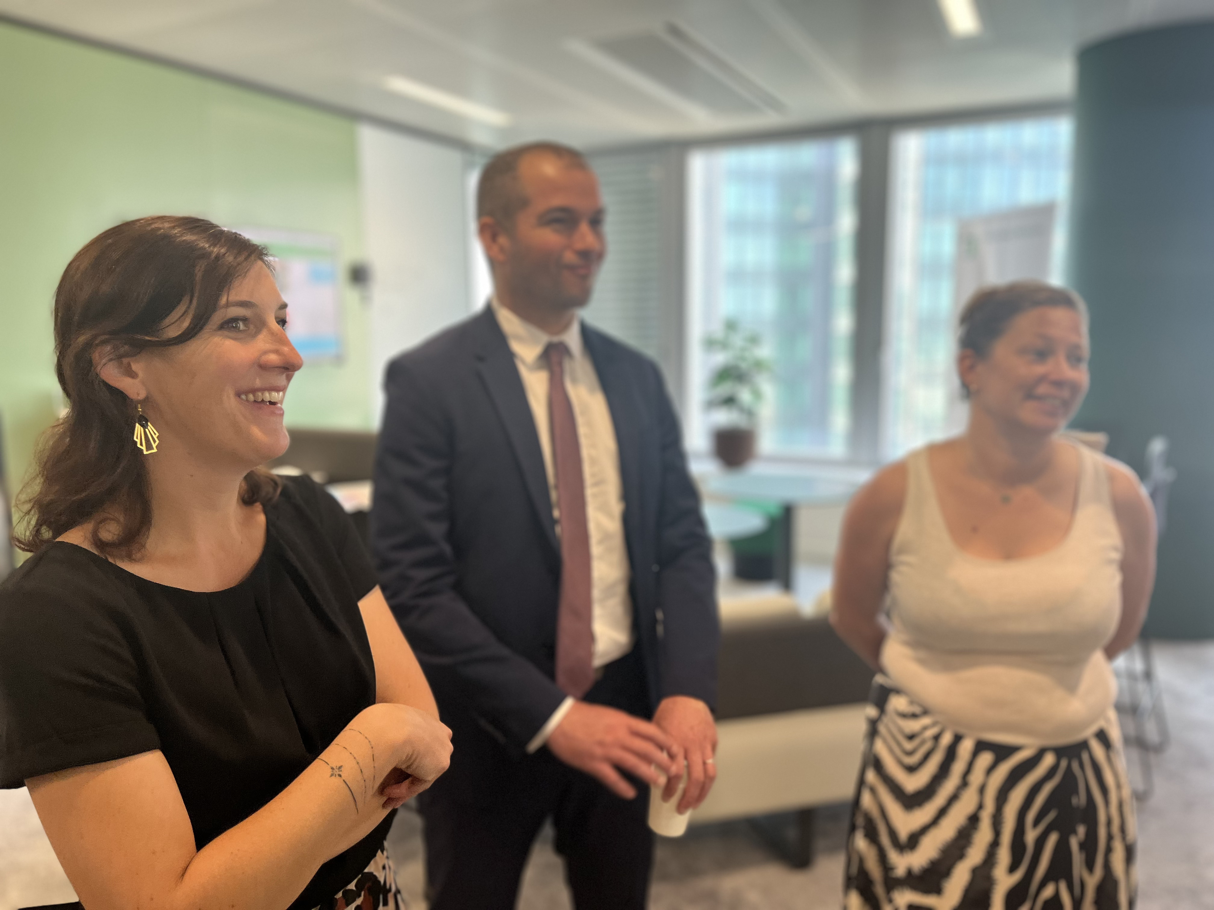 Trois personnes debout dans un espace de bureau moderne, discutant entre elles. Un homme en costume tient un gobelet au centre, encadré par deux femmes, l'une en robe noire à gauche et l'autre en haut blanc et jupe à motifs à droite. Fond avec grandes fenêtres offrant une vue sur des bâtiments extérieurs.