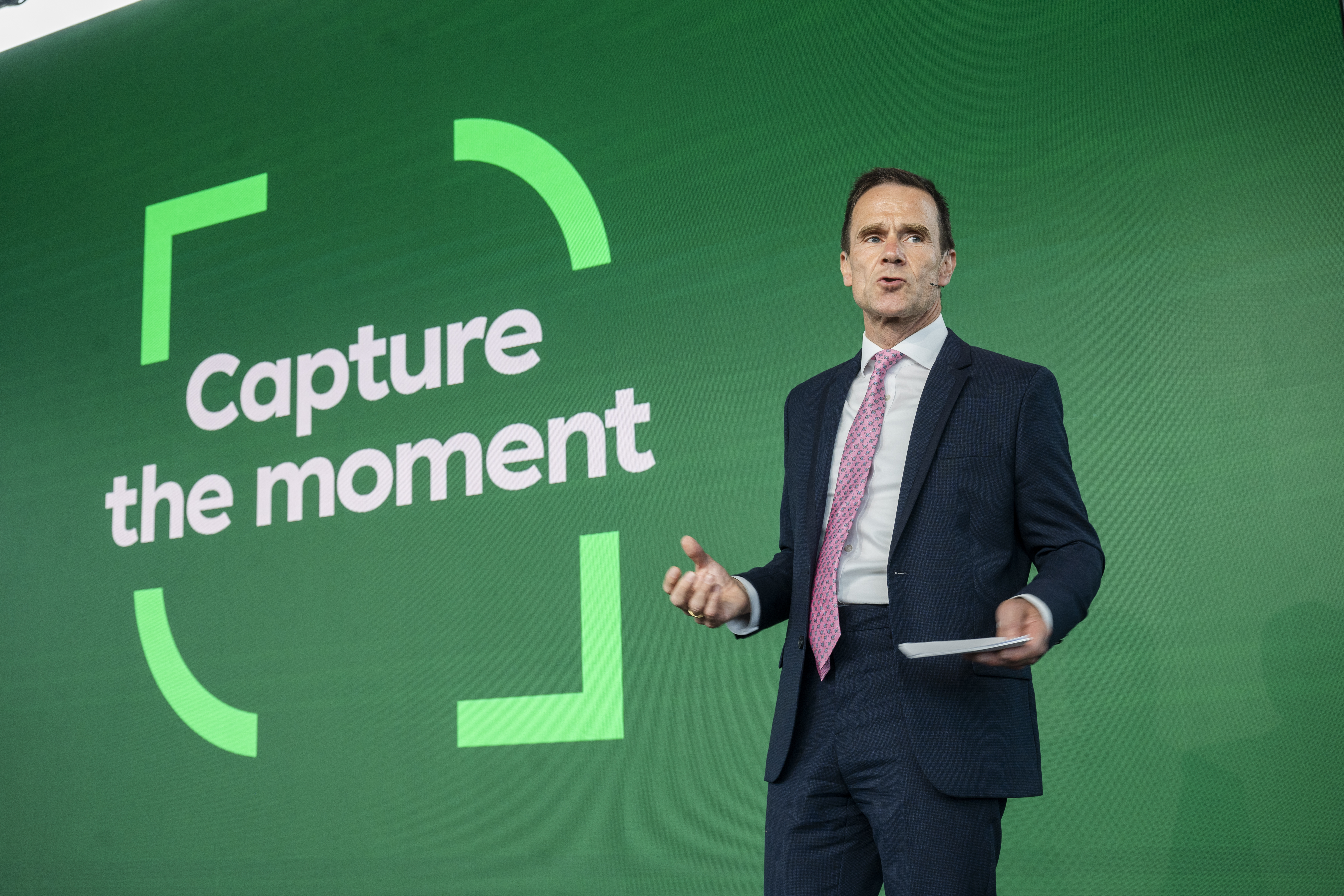 Un homme en costume sombre et cravate rose parle sur scène avec un fond vert affichant le texte "Capture the moment" encadré par des parenthèses stylisées.