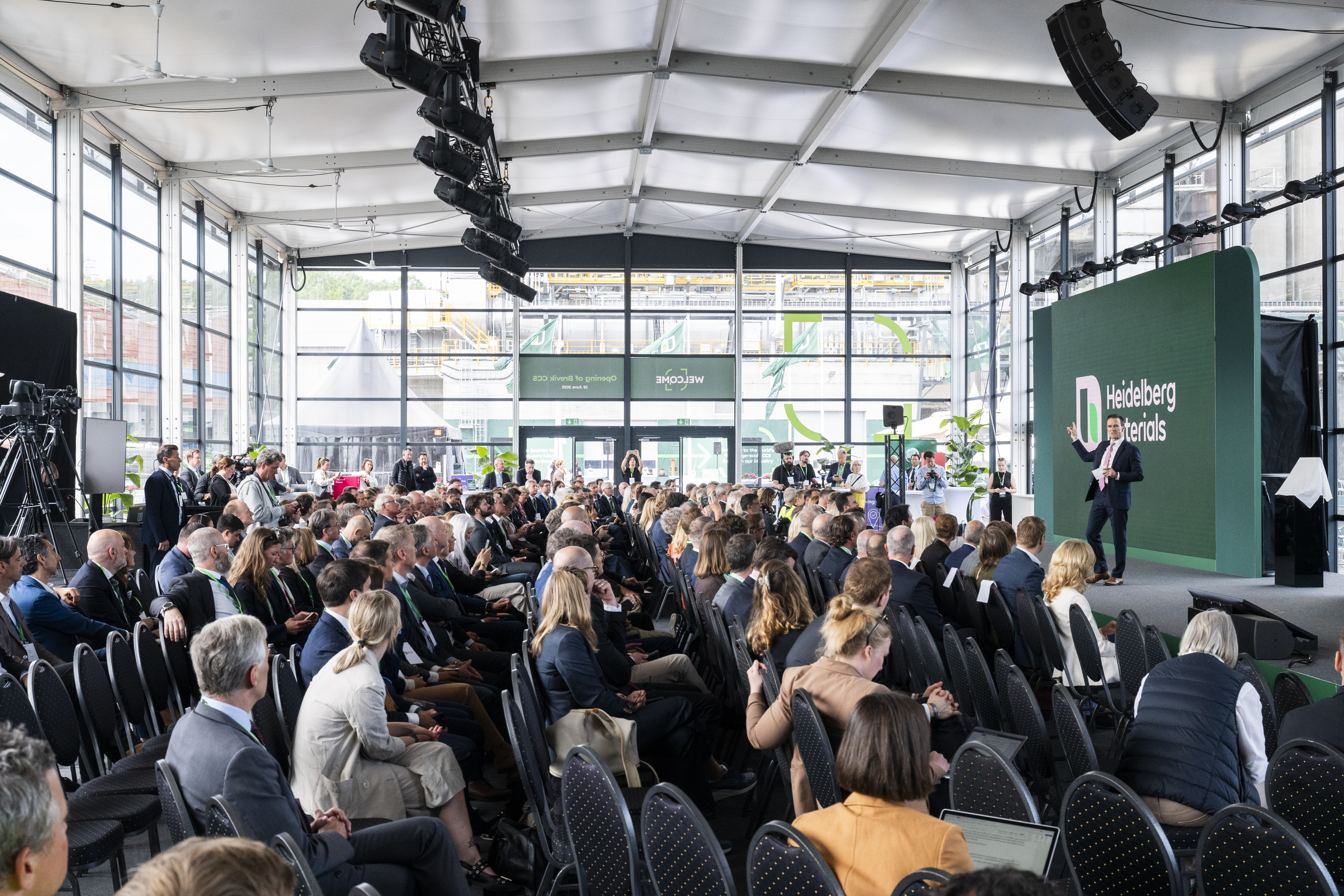 Salle de conférence lumineuse avec de grandes baies vitrées, remplie de participants assis face à une scène. Un conférencier en costume se tient à droite sur la scène, où figure un écran vert avec le logo et le texte "Heidelberg Materials". Des structures d'éclairage suspendues au plafond et des plantes disposées près des fenêtres.