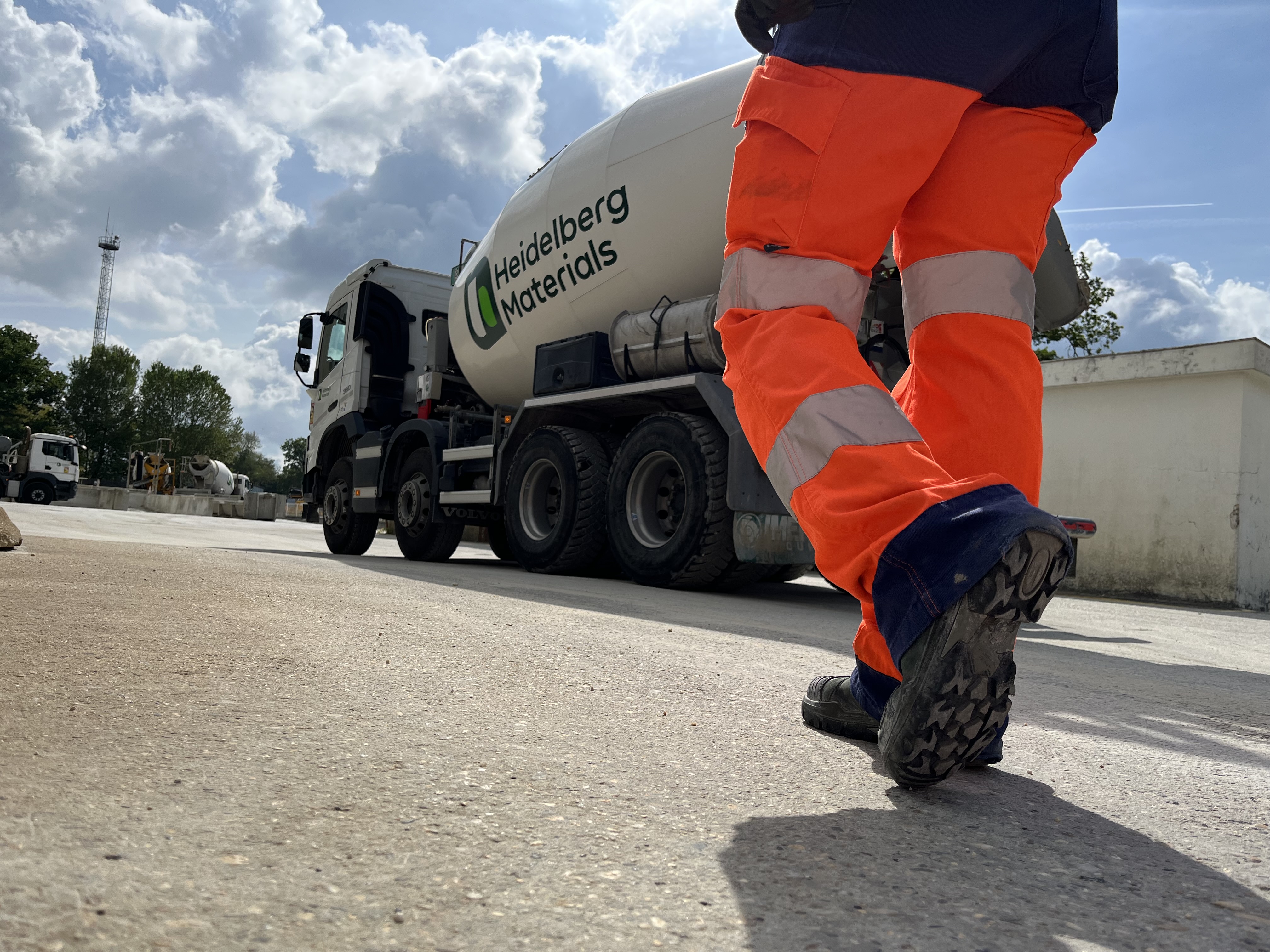 Un travailleur en pantalon orange fluorescent marche sur un site industriel, vu de dos et en contre-plongée. En arrière-plan, un camion-toupie portant le logo "Heidelberg Materials" stationne sous un ciel partiellement nuageux.