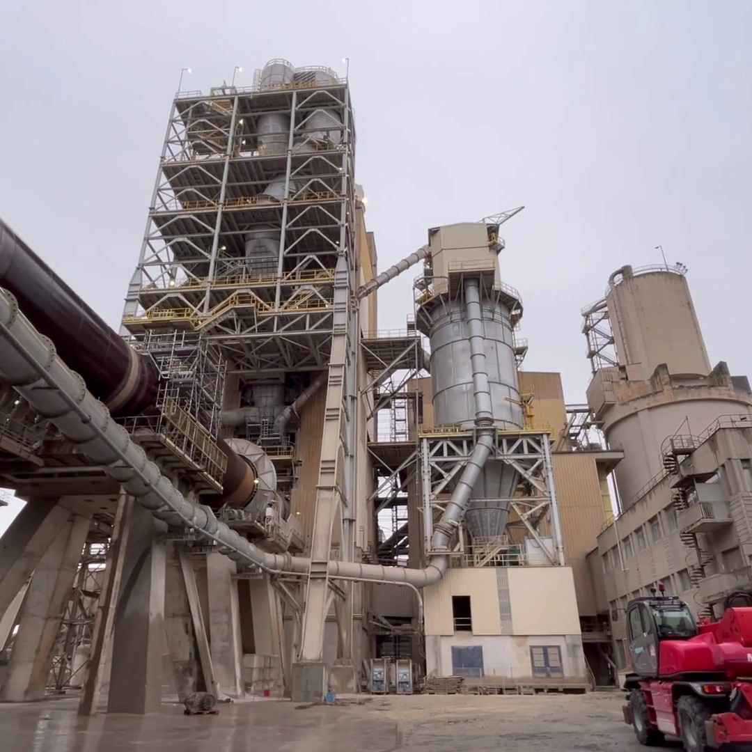 Installation industrielle avec de hautes structures métalliques et des tours cylindriques, reliées par de grands tuyaux et des échafaudages. Un véhicule de chantier rouge est garé au premier plan en bas à droite. Ciel couvert en arrière-plan.