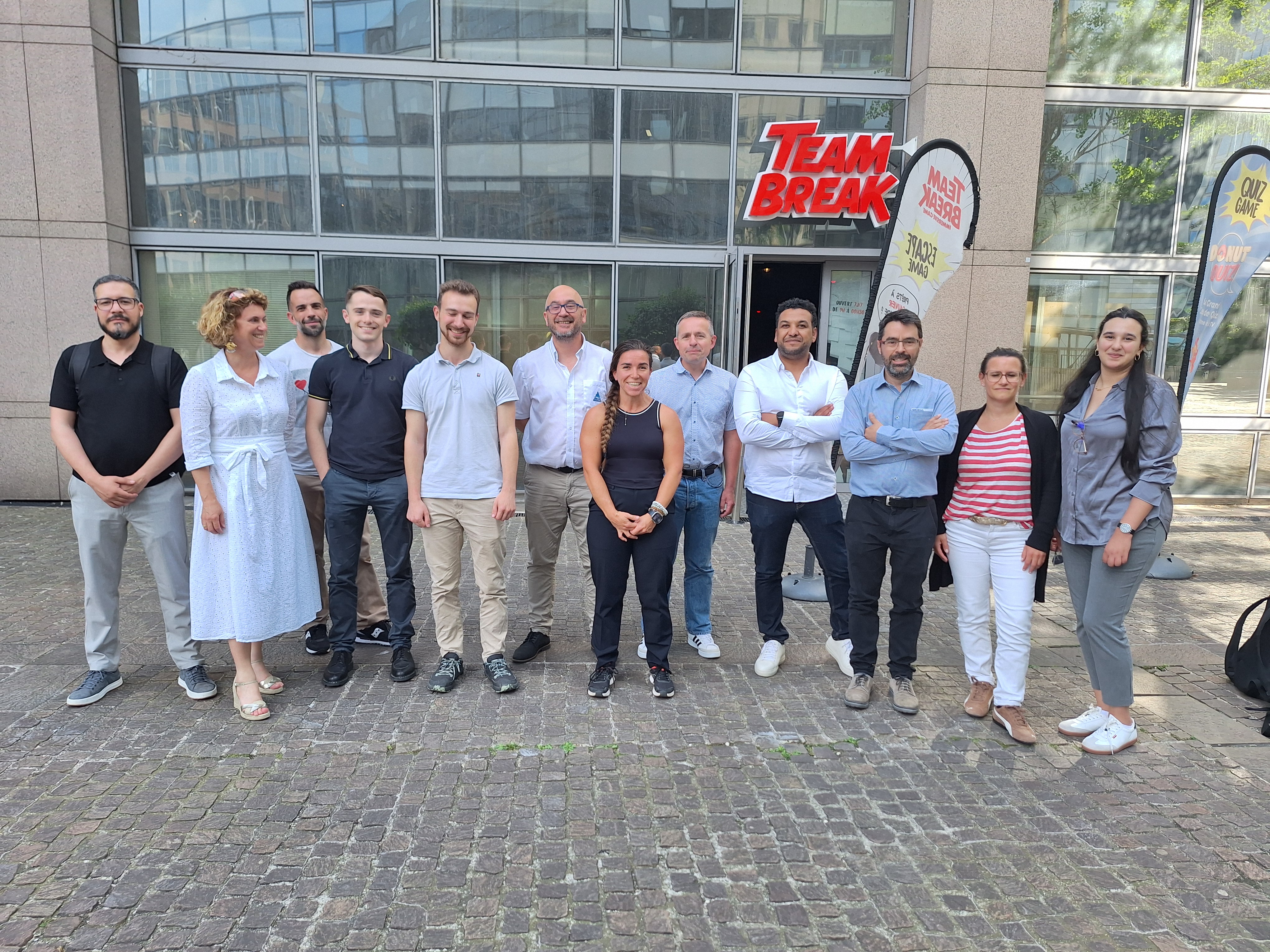 Un groupe de 12 personnes debout devant un bâtiment vitré avec une enseigne rouge « Team Break ». Deux drapeaux publicitaires à droite affichent le logo de la même enseigne. Le groupe est composé d’hommes et de femmes souriants, habillés en vêtements décontractés, posant sur un pavé extérieur par temps ensoleillé.