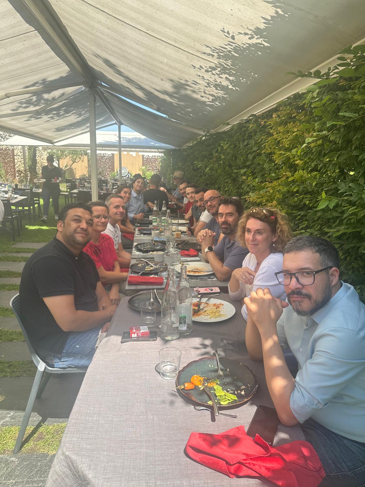 Groupe de personnes assises autour d'une longue table en extérieur, partageant un repas sous une pergola ombragée près d'un mur de verdure.