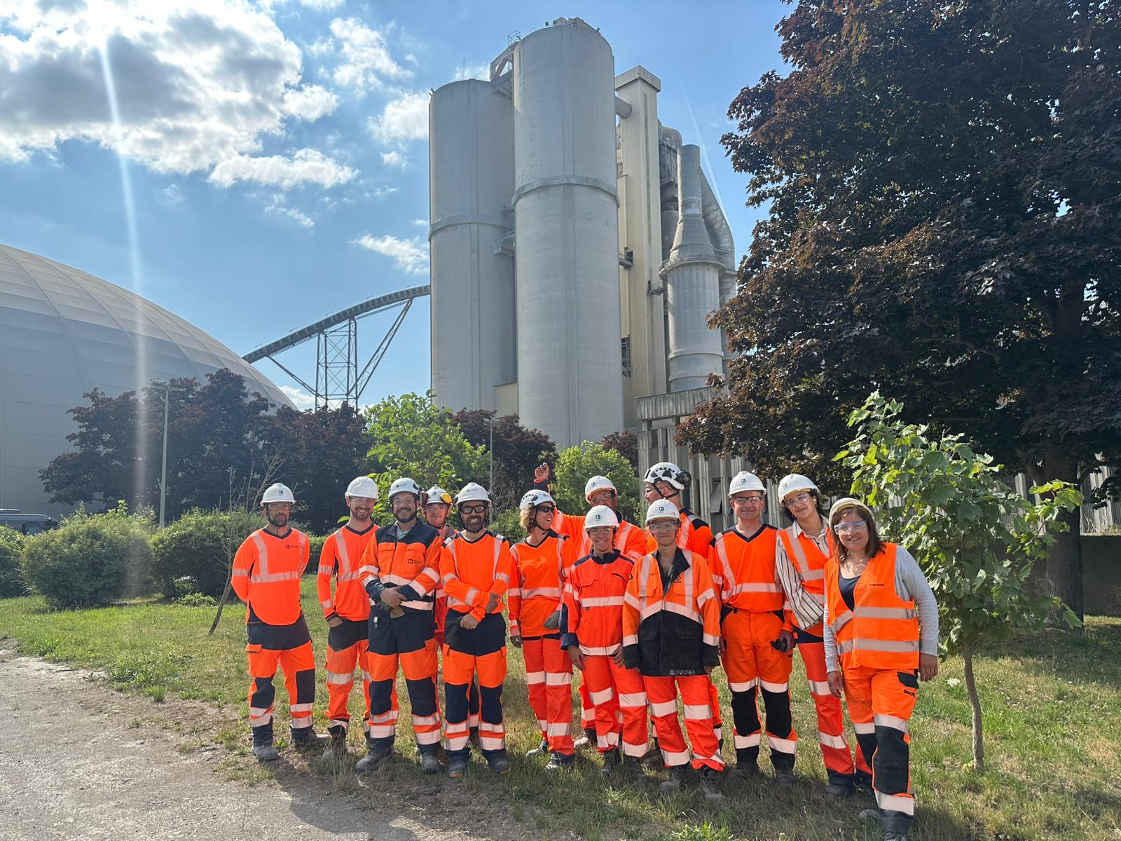 Groupe de treize personnes portant des équipements de sécurité orange et des casques blancs, debout devant une usine avec des silos industriels et un dôme en arrière-plan, entourés d'arbres et de pelouse par temps ensoleillé.