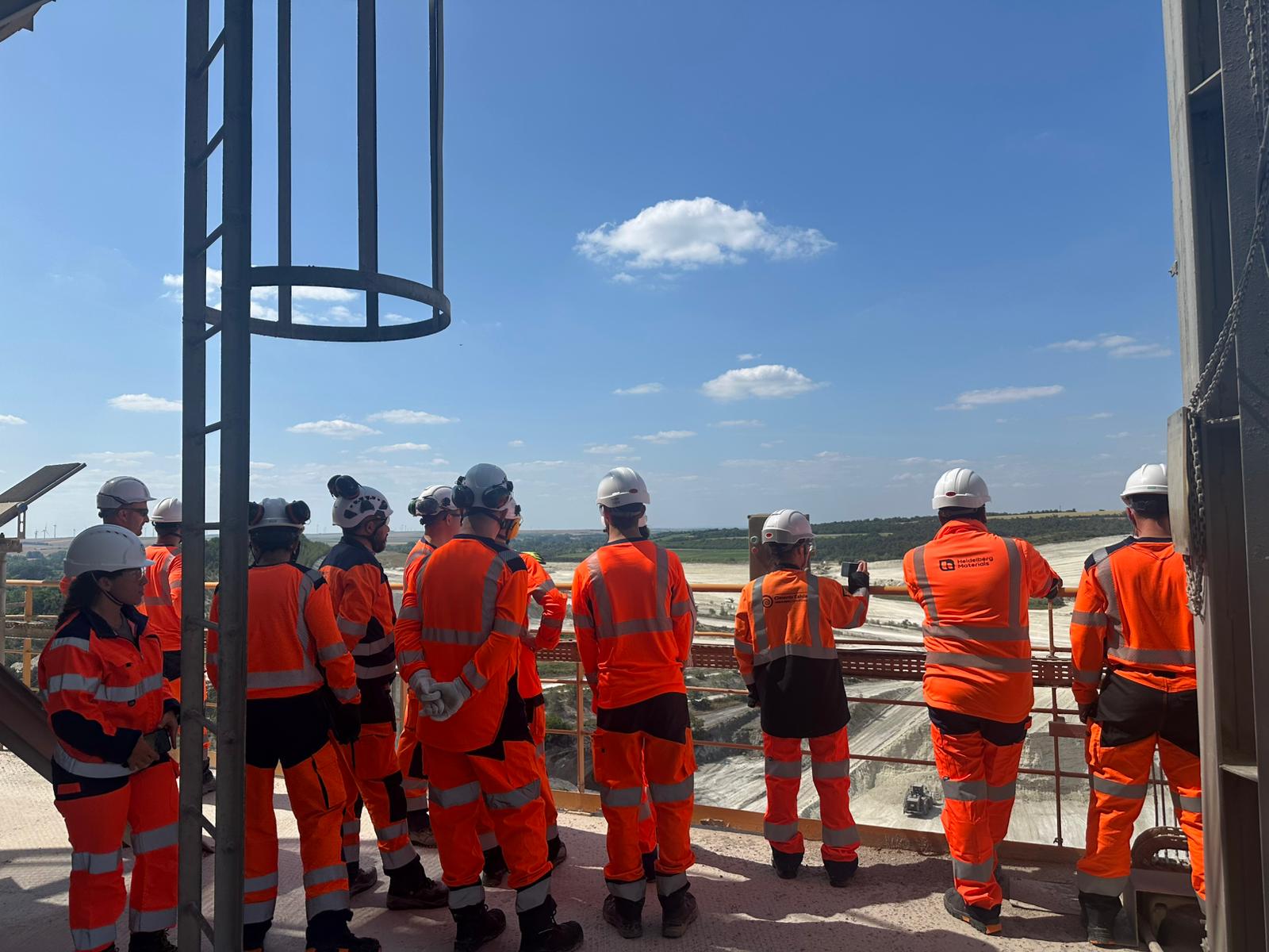 Un groupe de personnes en tenue de sécurité orange et casques blancs observe un chantier en extérieur depuis une plateforme surélevée par une journée ensoleillée, avec un paysage ouvert et un ciel bleu en arrière-plan.