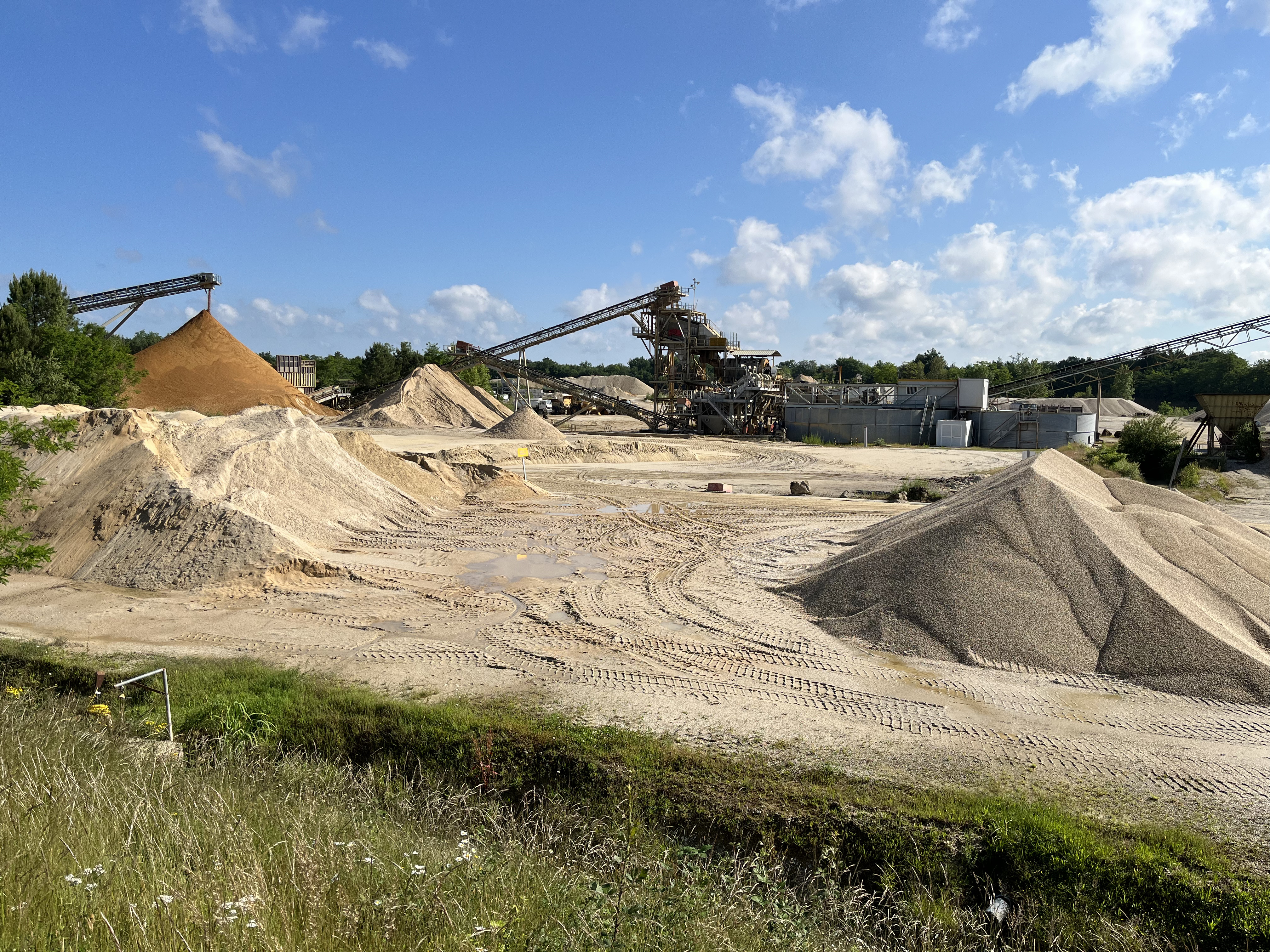 Site industriel de carrières sous un ciel bleu avec nuages. Tas de sable et de gravier au premier plan, convoyeurs inclinés transportant des matériaux vers des structures en arrière-plan, entourés de végétation.