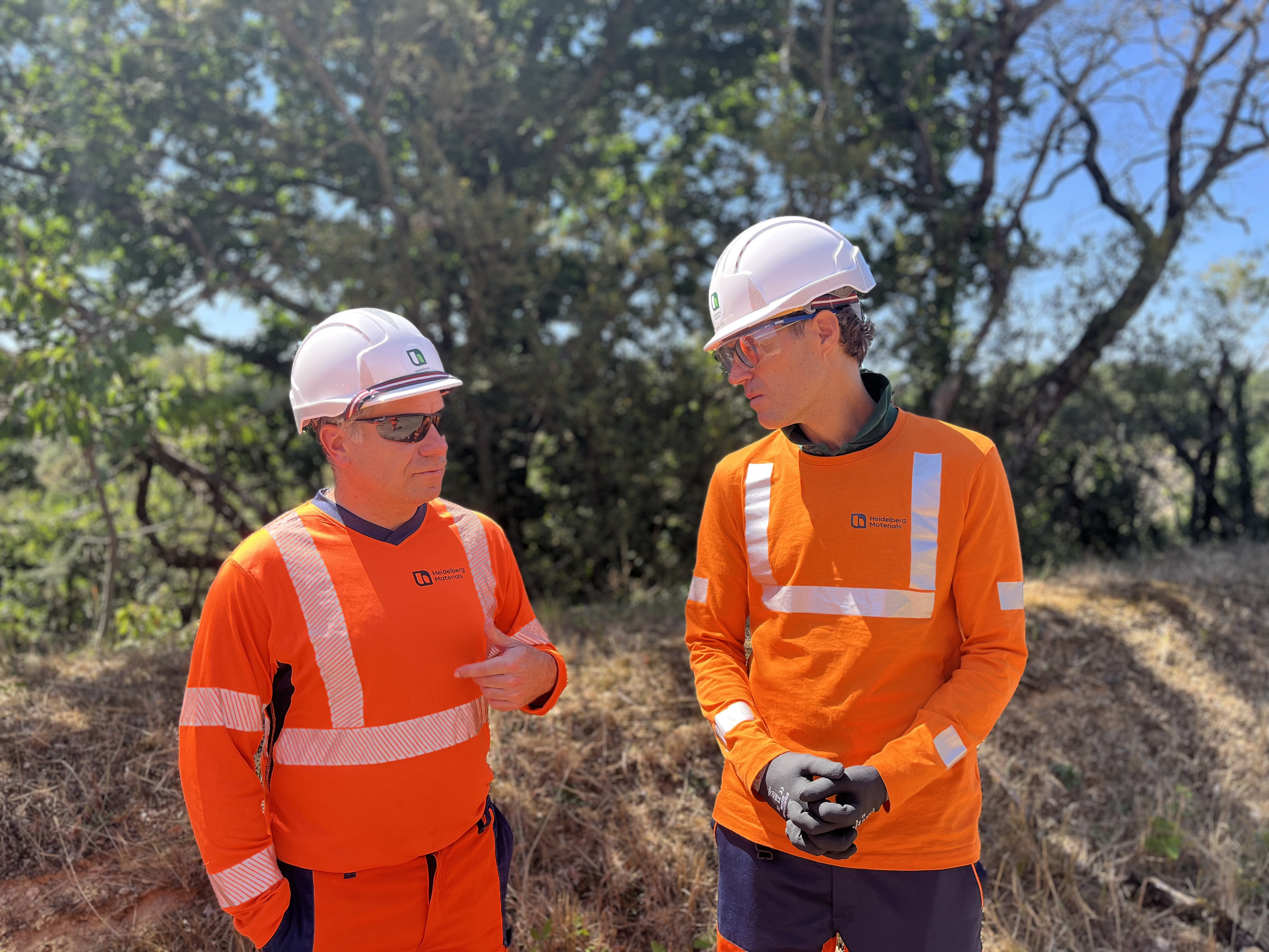 Deux personnes en tenue de sécurité orange avec casques blancs discutent à l'extérieur près d'une zone arborée, sous un ciel ensoleillé. L'une pointe du doigt en parlant tandis que l'autre écoute attentivement avec des gants noirs.