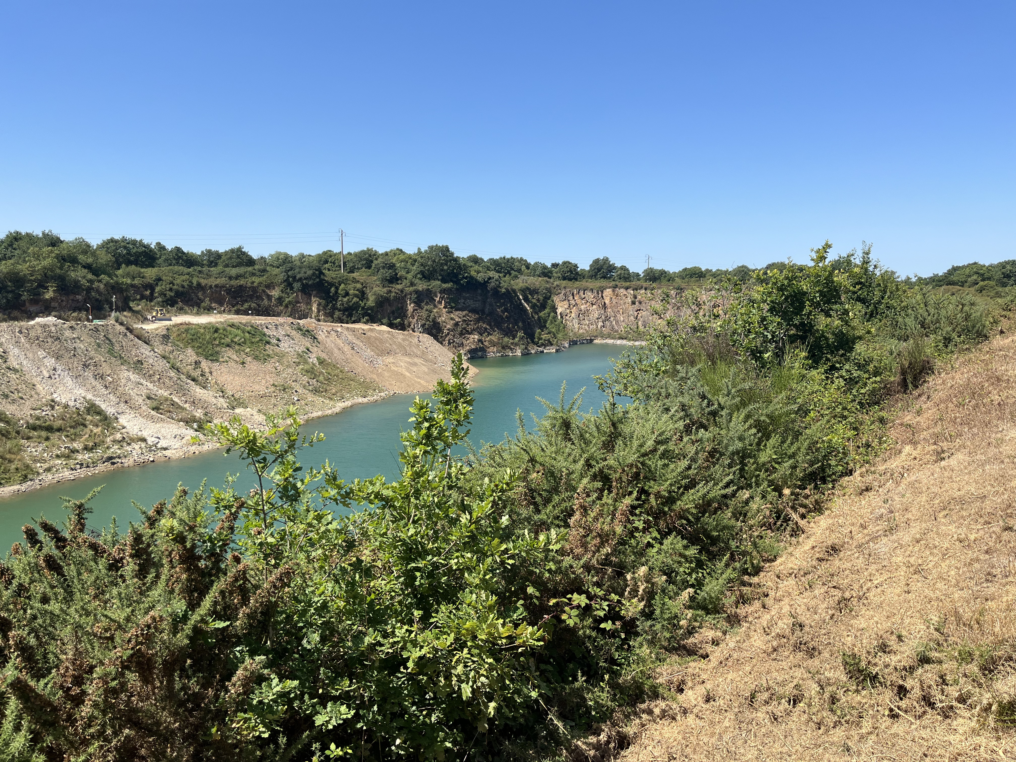 Vue d'une ancienne carrière transformée en lac, entourée de végétation dense. L'eau turquoise est bordée de falaises abruptes sur la gauche et de buissons sur la droite, sous un ciel bleu clair.