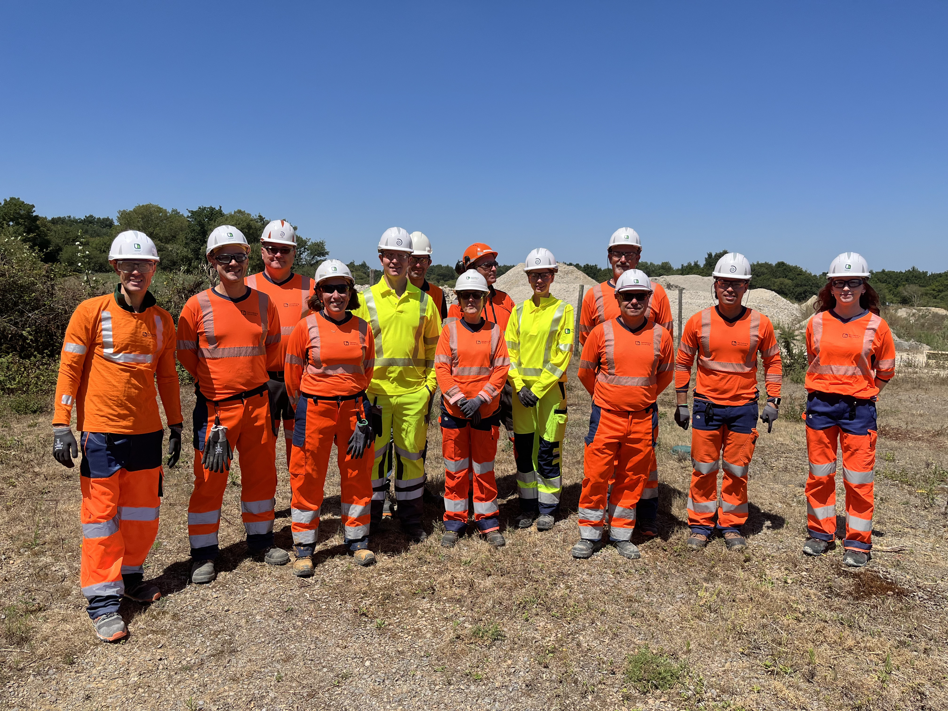 Groupe de 13 travailleurs en tenue de sécurité orange et jaune avec casques, debout à l'extérieur sur un terrain dégagé par une journée ensoleillée, avec des arbres et des collines en arrière-plan.