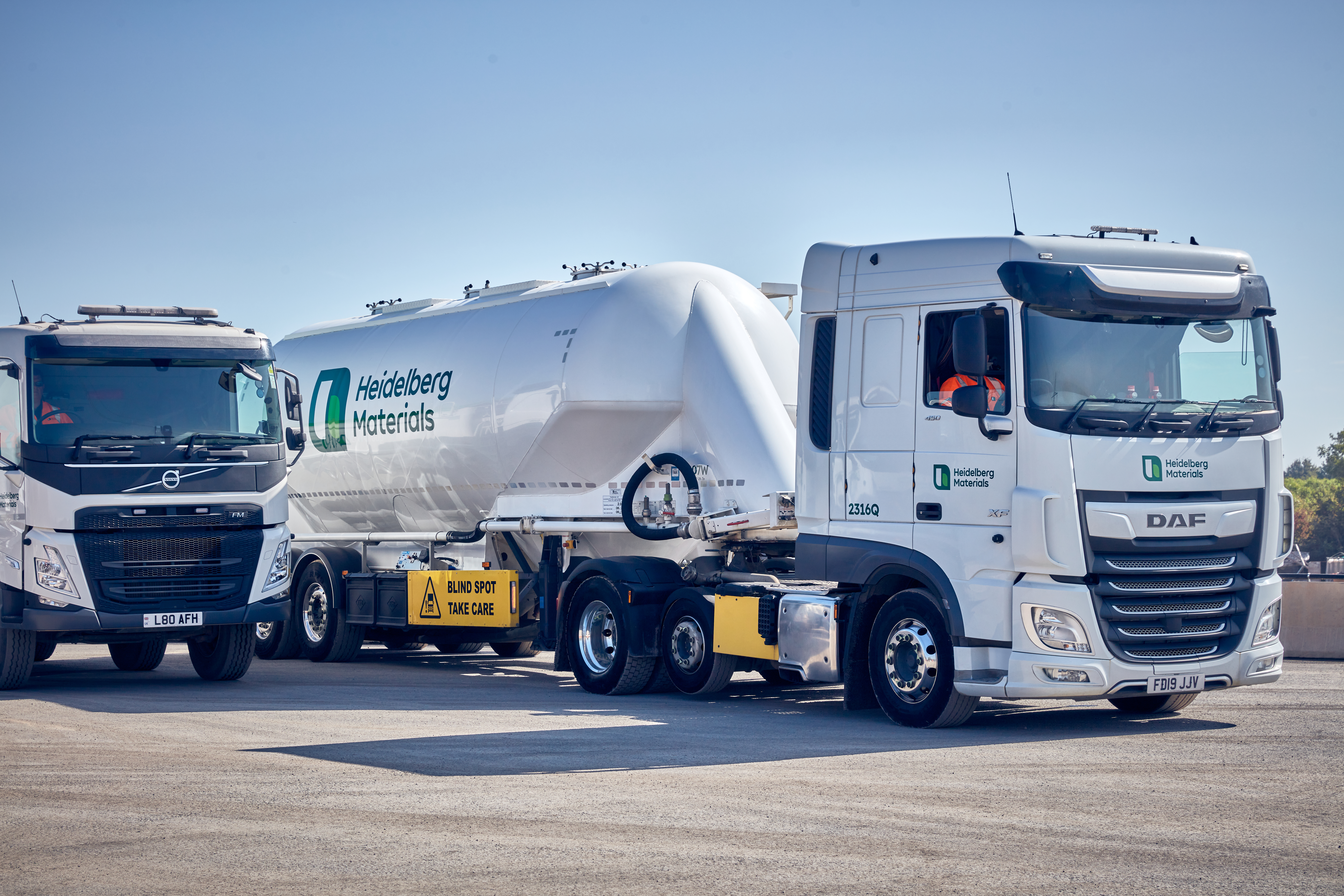 Deux camions blancs de transport routier, l’un à gauche partiellement visible et l’autre complètement à droite, stationnés sur une aire dégagée. Le camion de droite porte une citerne blanche avec le logo "Heidelberg Materials" bien visible. Ciel bleu clair en arrière-plan.