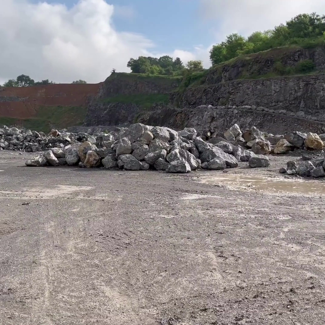 Tas de rochers gris au centre d'une carrière ouverte avec un sol terreux et des parois rocheuses en arrière-plan, entouré de collines verdoyantes sous un ciel partiellement nuageux.