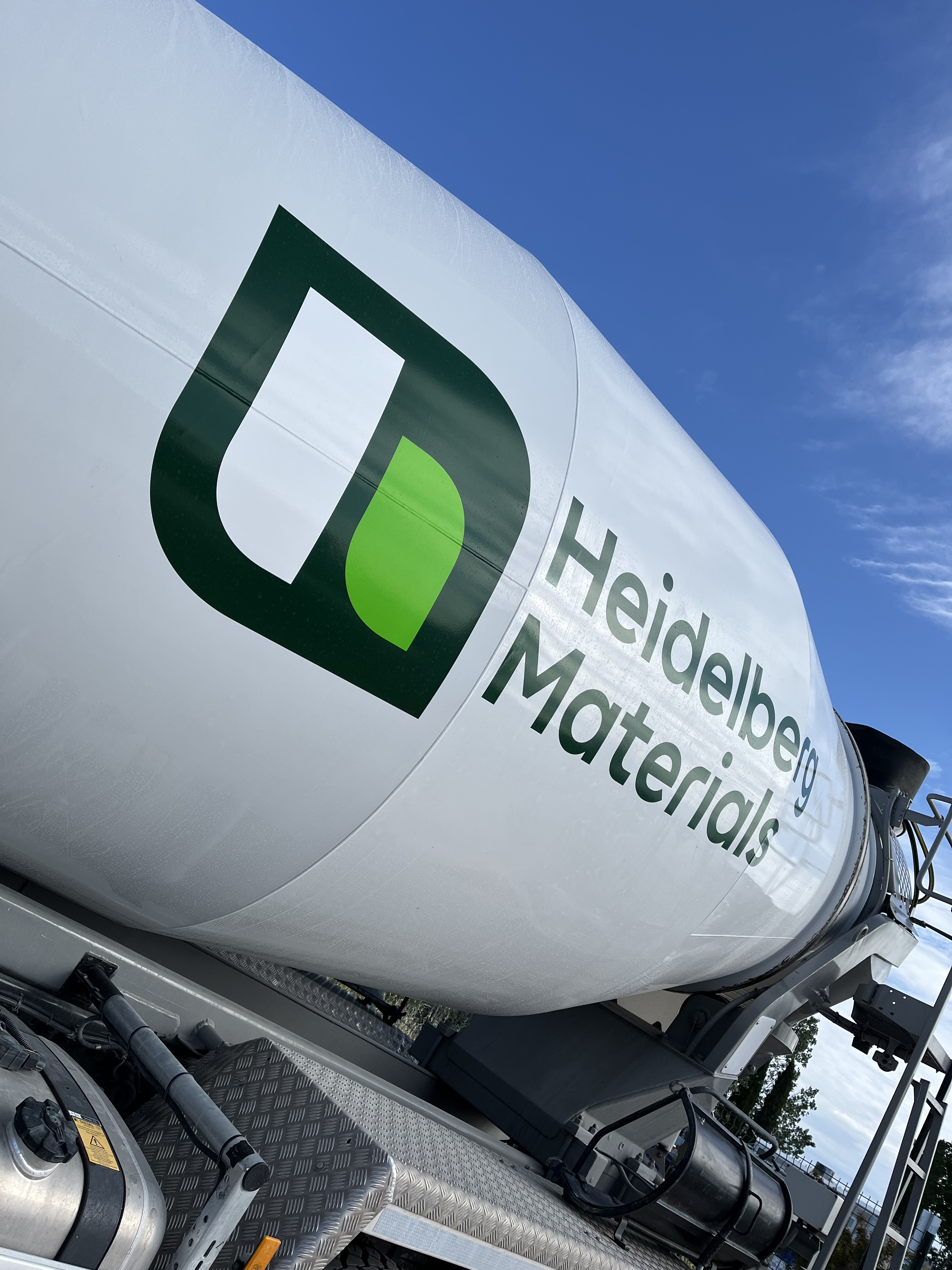 Camion toupie avec un tambour blanc affichant le logo et le nom "Heidelberg Materials" en vert et noir, sur fond de ciel bleu.