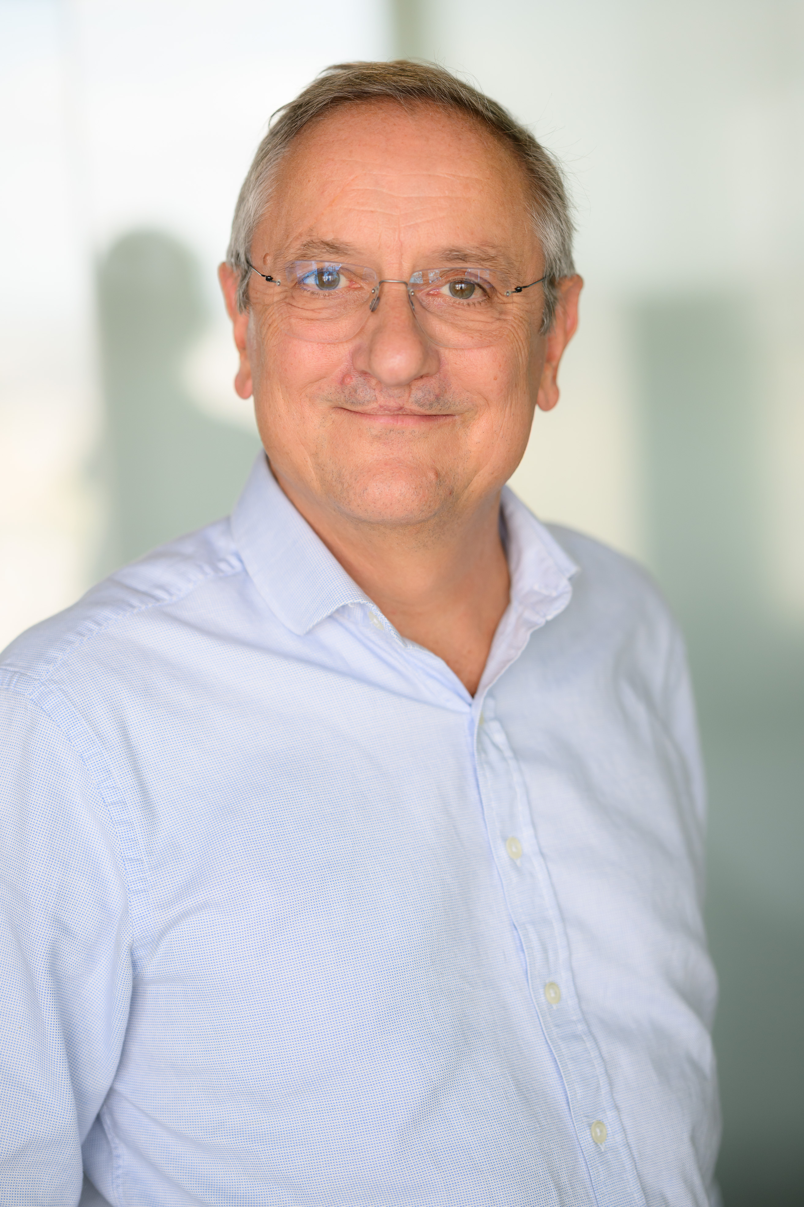 A person wearing a light blue button-up shirt, standing in front of a blurred office background with glass panels.