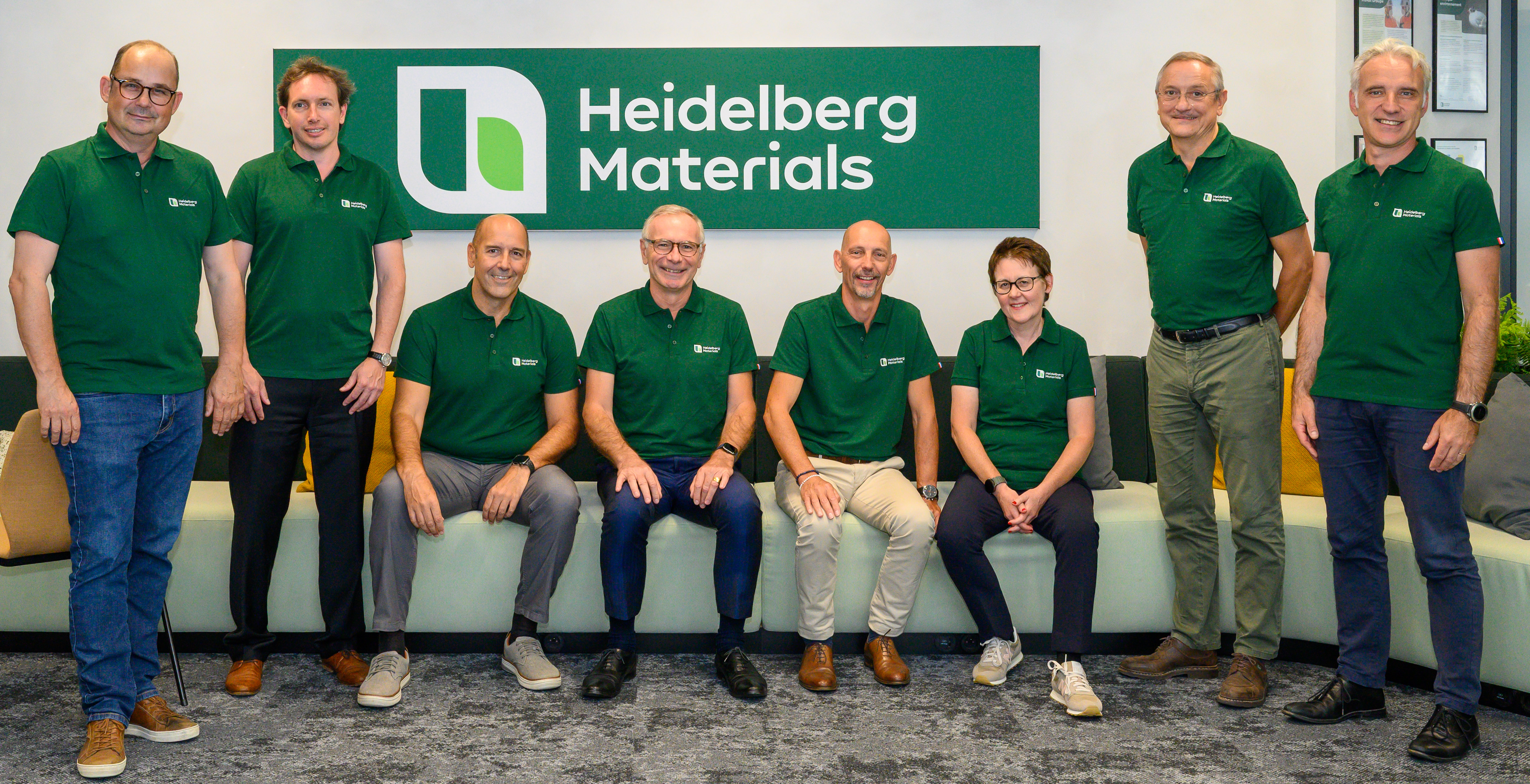 Huit personnes en polo vert avec le logo "Heidelberg Materials" posent devant un panneau de l'entreprise. Quatre sont debout à l'arrière et quatre assises sur un banc devant, formant deux rangées. L'arrière-plan inclut un mur blanc et une moquette grise.