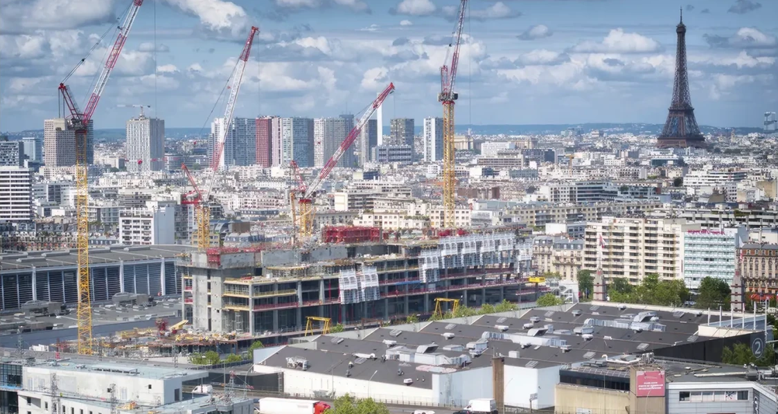 Vue panoramique d'une zone urbaine en construction à Paris, avec plusieurs grues rouges et jaunes entourant des bâtiments en développement au centre. En arrière-plan, plusieurs immeubles modernes et la tour Eiffel visible à droite sous un ciel partiellement nuageux.