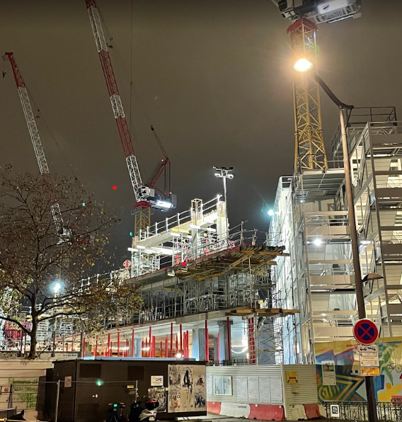 Chantier de construction nocturne avec plusieurs grues illuminées et un immeuble en cours de construction à droite. Des panneaux de signalisation, barrières rouges et blanches, et une zone éclairée par des lampes de chantier occupent le premier plan. Un arbre sans feuilles se trouve à gauche.