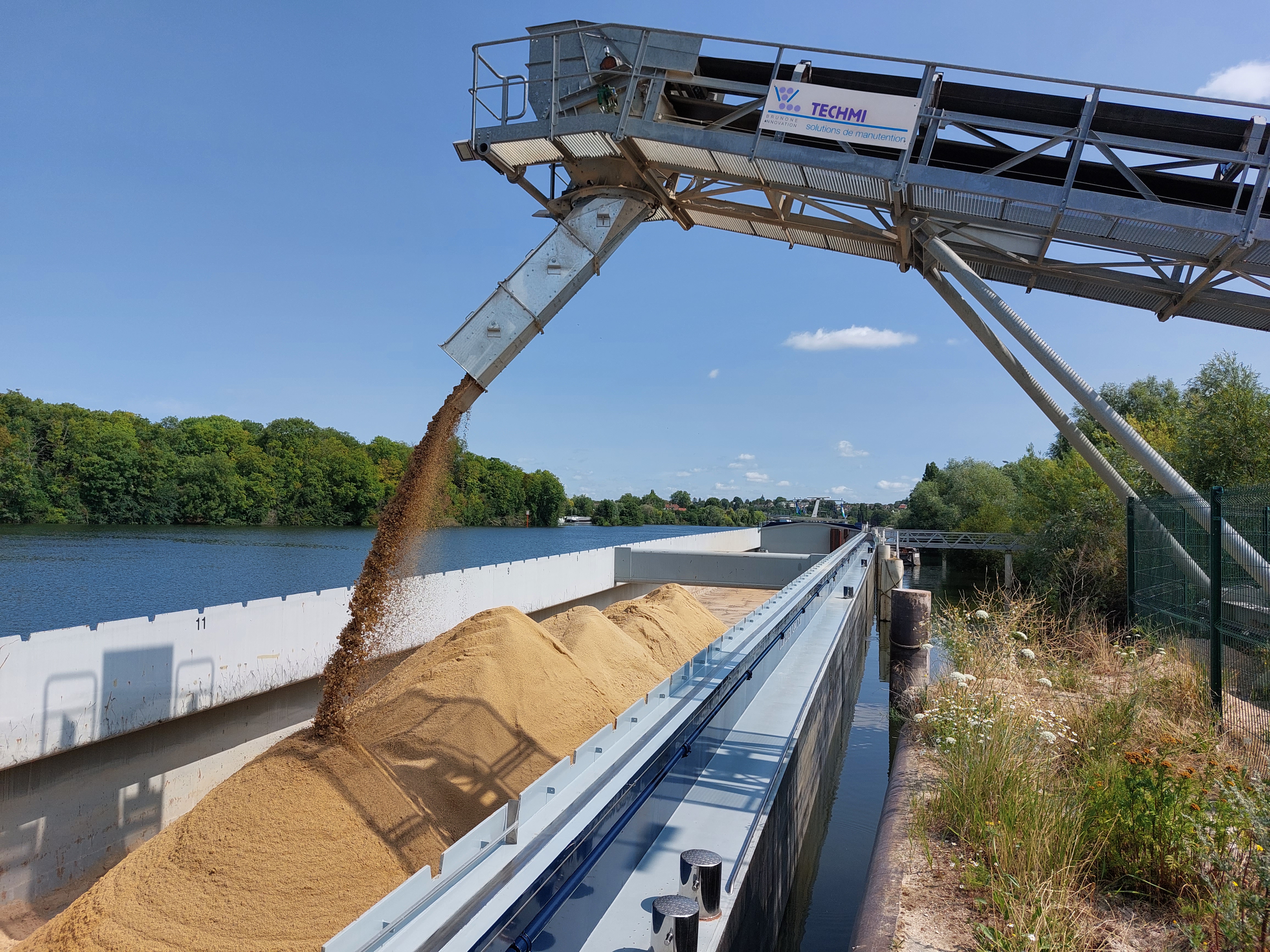 Sable déversé d'un convoyeur dans une péniche stationnée sur un cours d'eau, entouré de végétation et d'arbres en arrière-plan sous un ciel bleu.