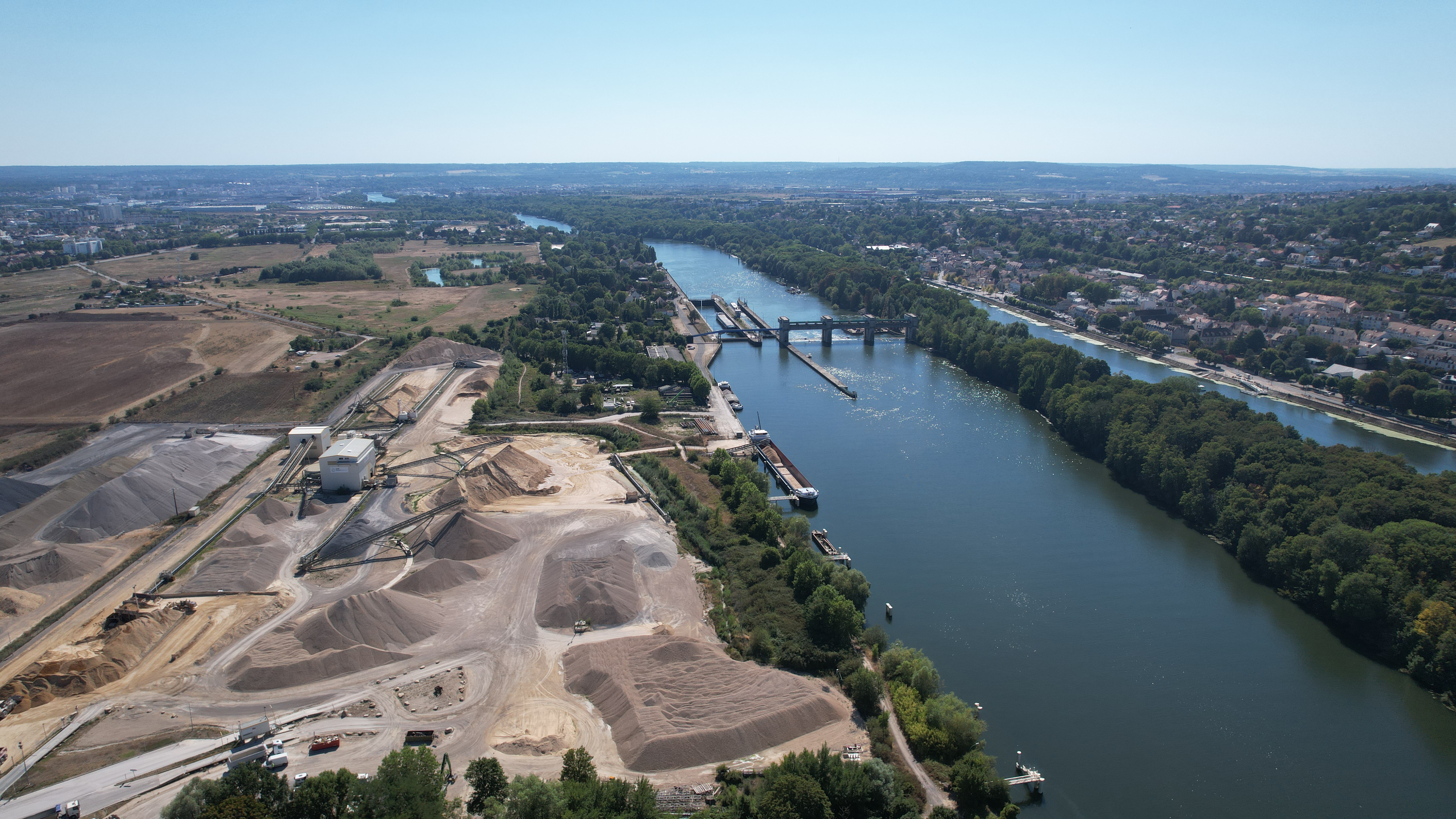 Vue aérienne d'une large rivière bordée d'arbres verts à droite et d'un site industriel avec de grands tas de gravier et d'équipements à gauche. Un pont enjambe la rivière au loin, reliant le paysage rural à gauche à une petite zone urbaine à droite sous un ciel bleu clair.