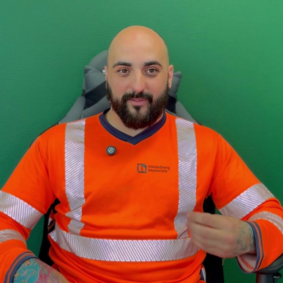 L'image montre un homme chauve avec une barbe, portant un pull orange vif à manches longues avec des bandes réfléchissantes blanches et le logo "Heidelberg Materials". Il est assis sur une chaise devant un fond vert uni. Il semble regarder directement la caméra, et sa posture suggère qu'il pourrait être en train de parler ou de faire une présentation vidéo. Son expression est neutre, voire légèrement souriante. Une partie d'un tatouage est visible sur son bras gauche.