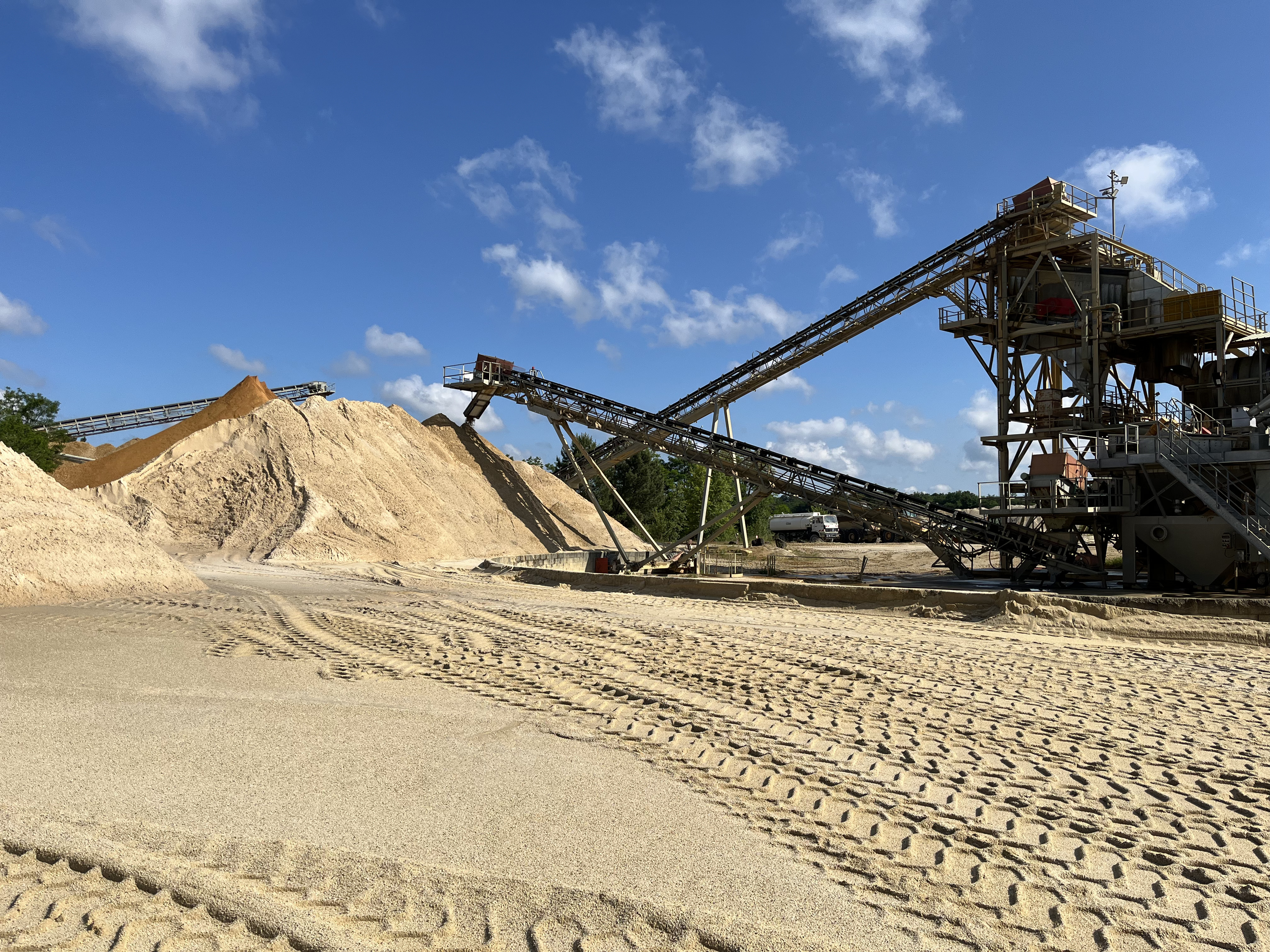Installation industrielle pour le traitement de sable, avec de grandes piles de sable à gauche et des convoyeurs mécaniques reliant des structures métalliques à droite. Sol avec des empreintes de pneus visibles au premier plan. Ciel bleu avec quelques nuages en arrière-plan.