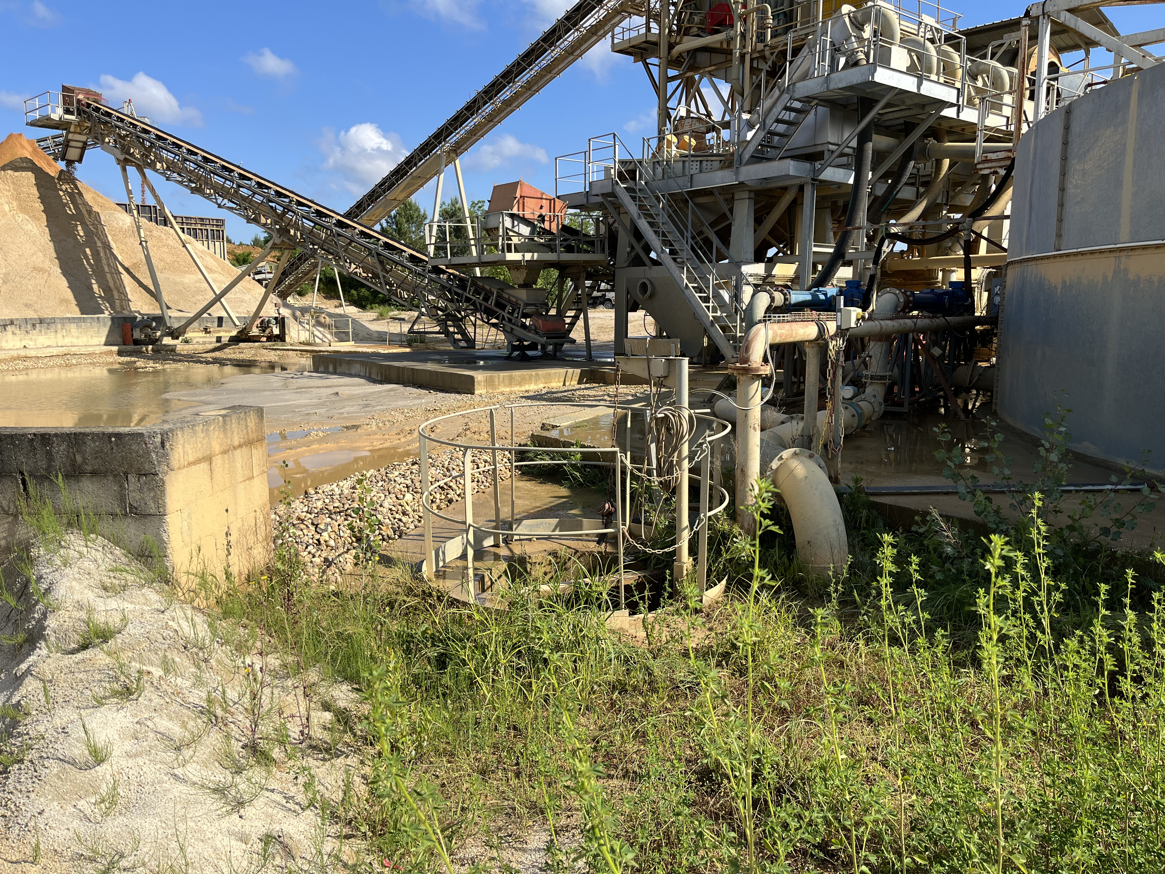 Machines industrielles sur un site de traitement du sable sous un ciel bleu clair, avec des bandes transporteuses acheminant le sable à gauche, un bassin de décantation à droite et une végétation luxuriante au premier plan.