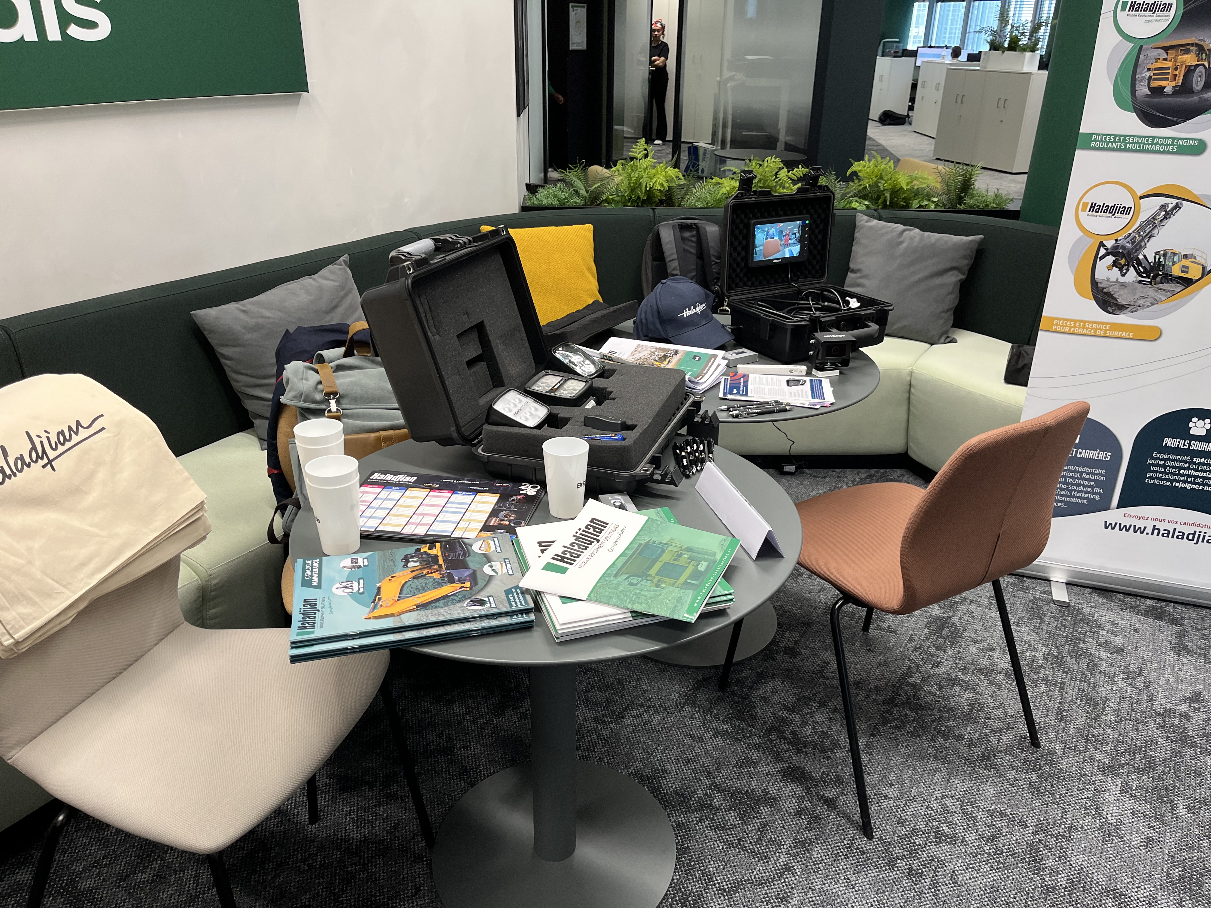 Table ronde dans une salle d'attente, présentant des équipements techniques divers, brochures, tasses et un écran de contrôle ouvert. En arrière-plan, canapé vert, affiches promotionnelles et plantes décoratives.