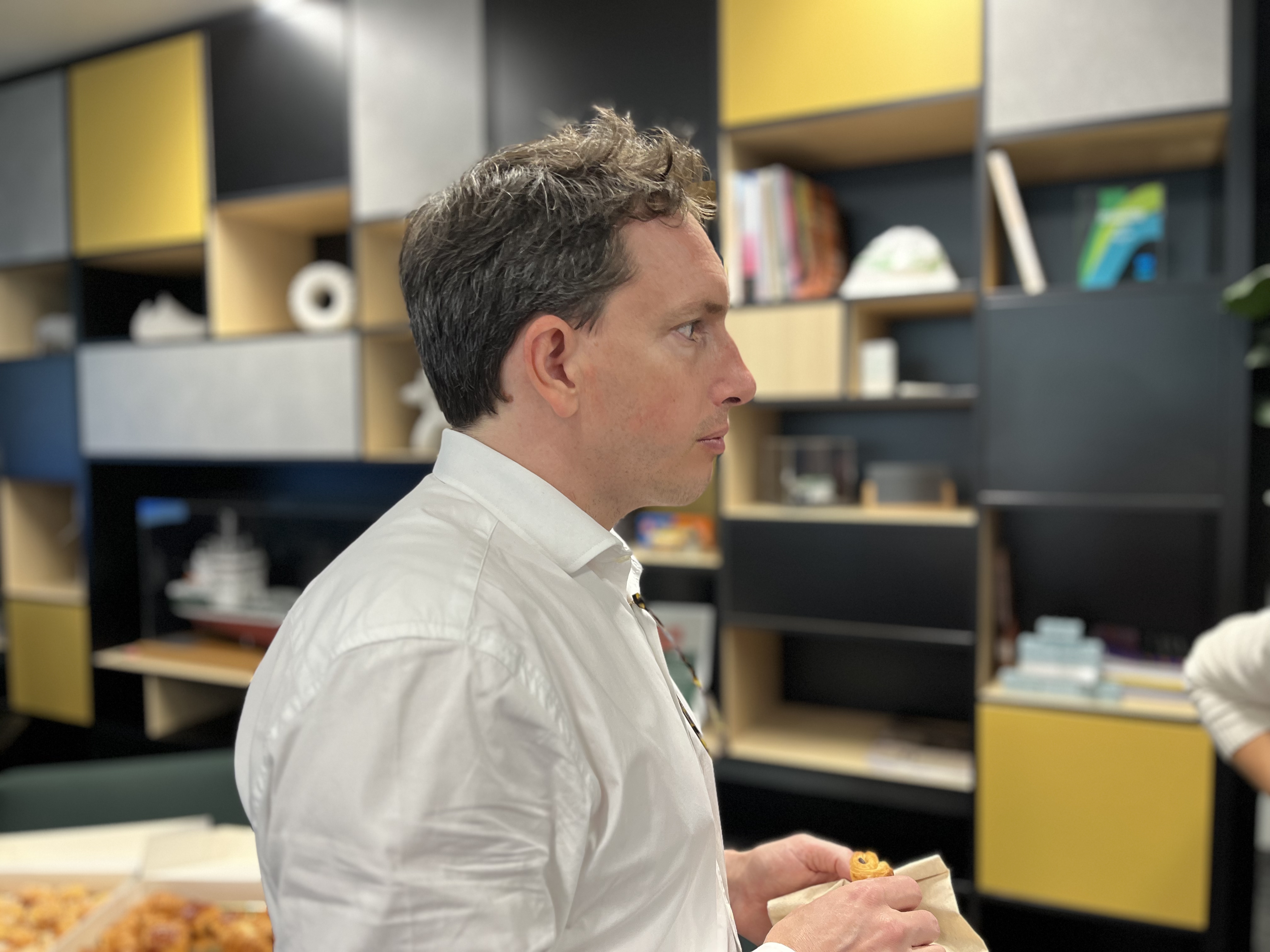 Un homme en chemise blanche, de profil, se tient dans un bureau moderne avec des étagères ouvertes noires et jaunes en arrière-plan. Sur une table au premier plan, des plateaux contiennent des aliments. Une personne partiellement visible est située à droite de l'image.