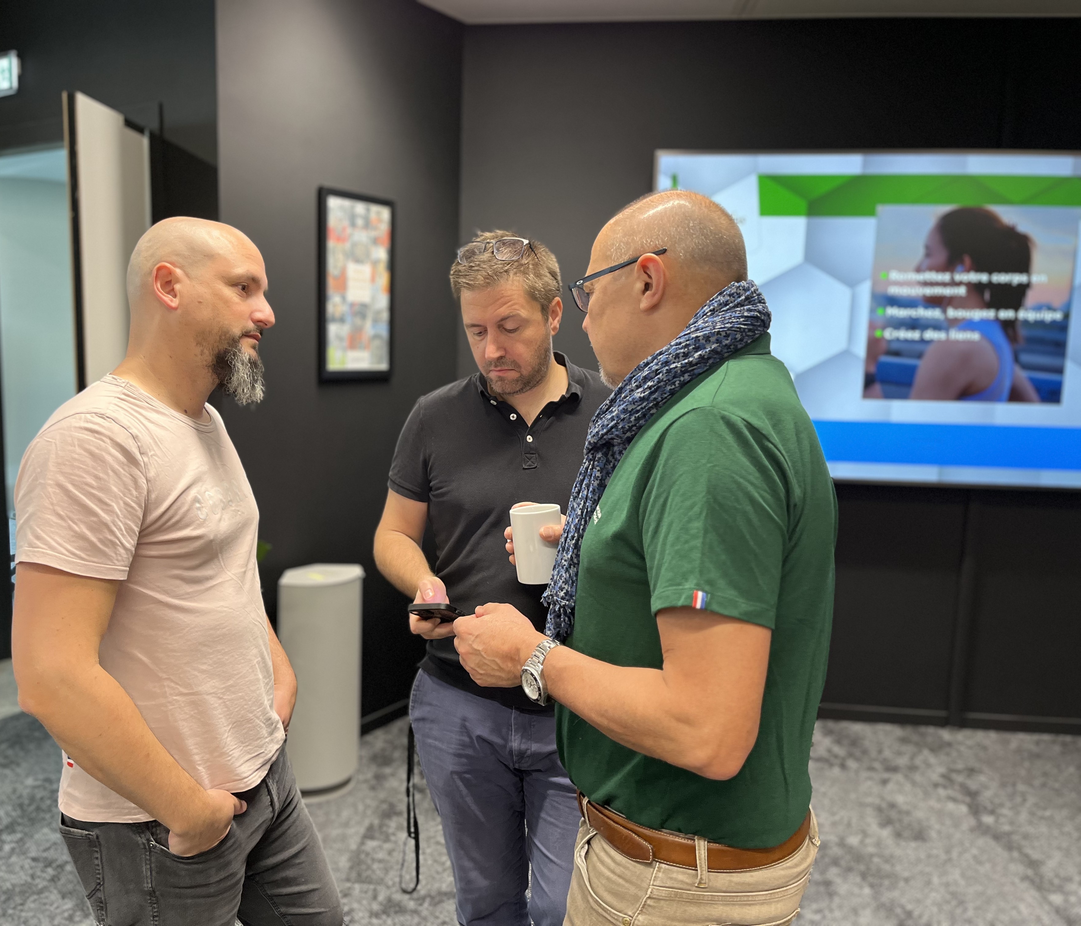 Trois hommes discutent debout dans un espace intérieur moderne. L'un tient une tasse de café et un autre regarde son téléphone. En arrière-plan, un écran affiche une présentation avec une image d’une femme et du texte. Une porte et un cadre sont visibles à gauche.