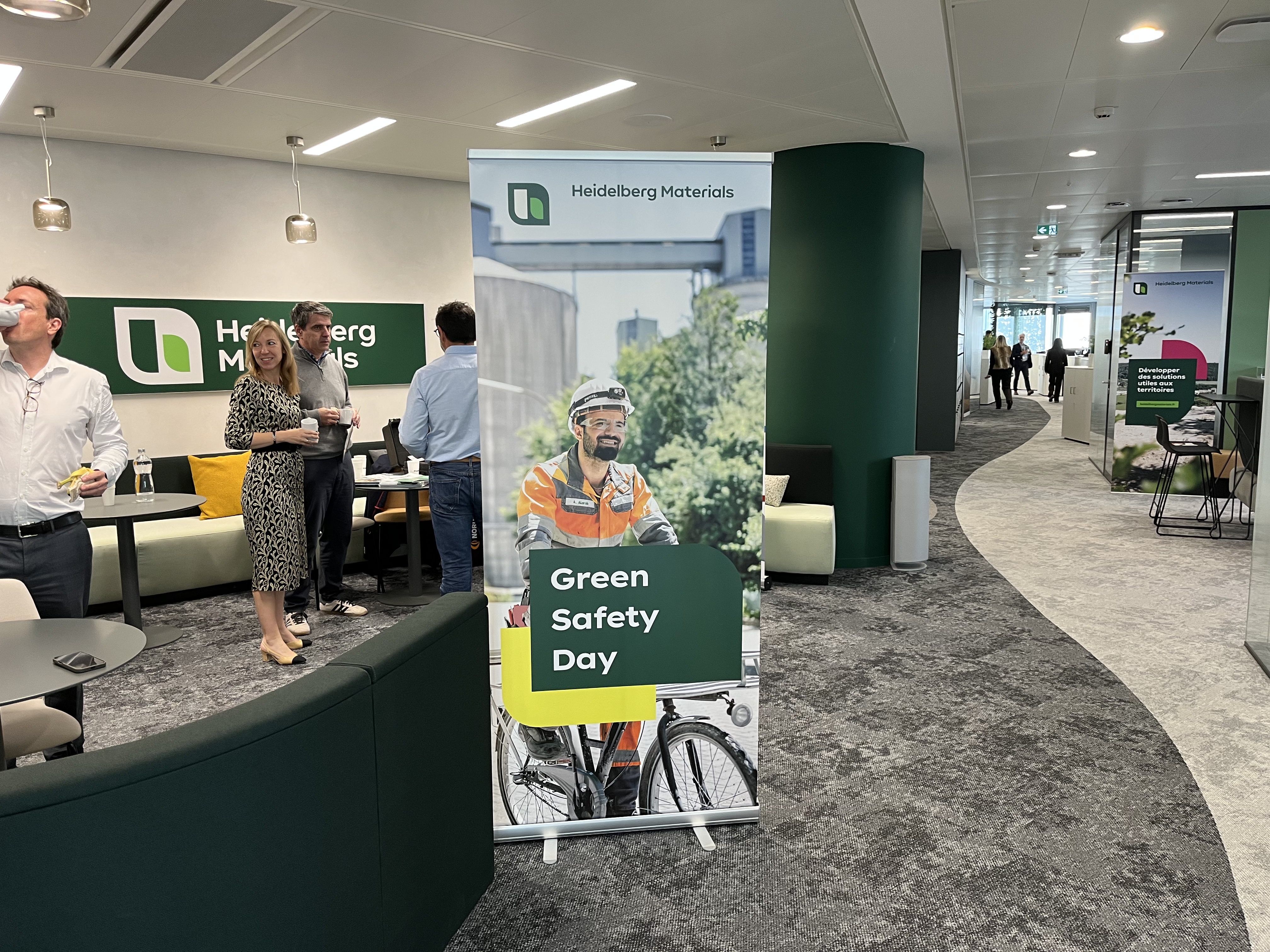 Bureau moderne avec des collaborateurs debout et discutant. Au centre, un panneau vertical affichant « Green Safety Day » avec un cycliste en uniforme orange et un fond de végétation. Mobilier design et zones de réunion visibles à l’arrière-plan.