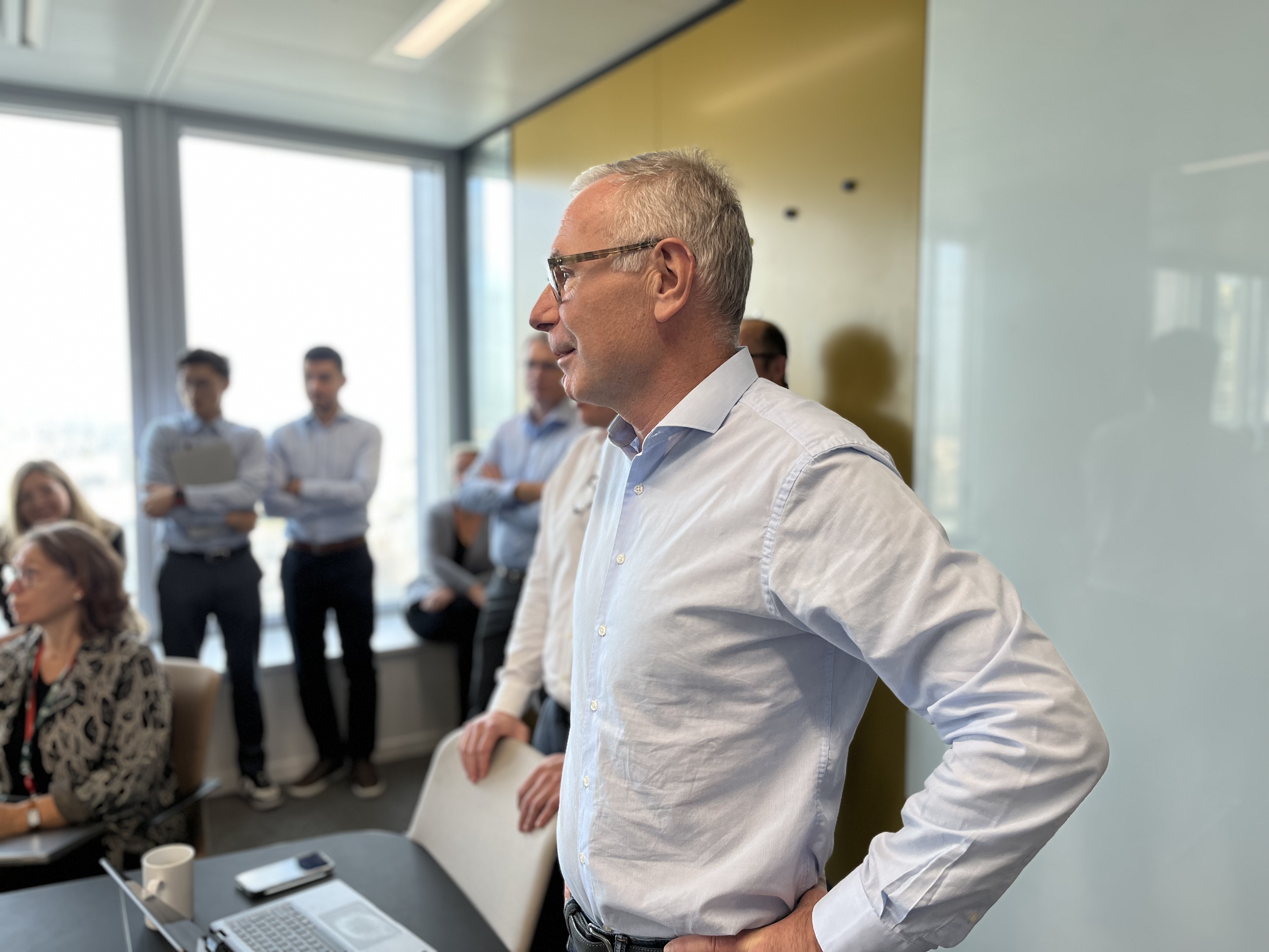 Un homme debout dans une salle de réunion moderne, entouré de collègues assis et debout. Une table au premier plan avec un ordinateur portable ouvert et des documents. Fenêtres lumineuses et mur jaune en arrière-plan. Scène d'une discussion professionnelle ou d'une présentation.