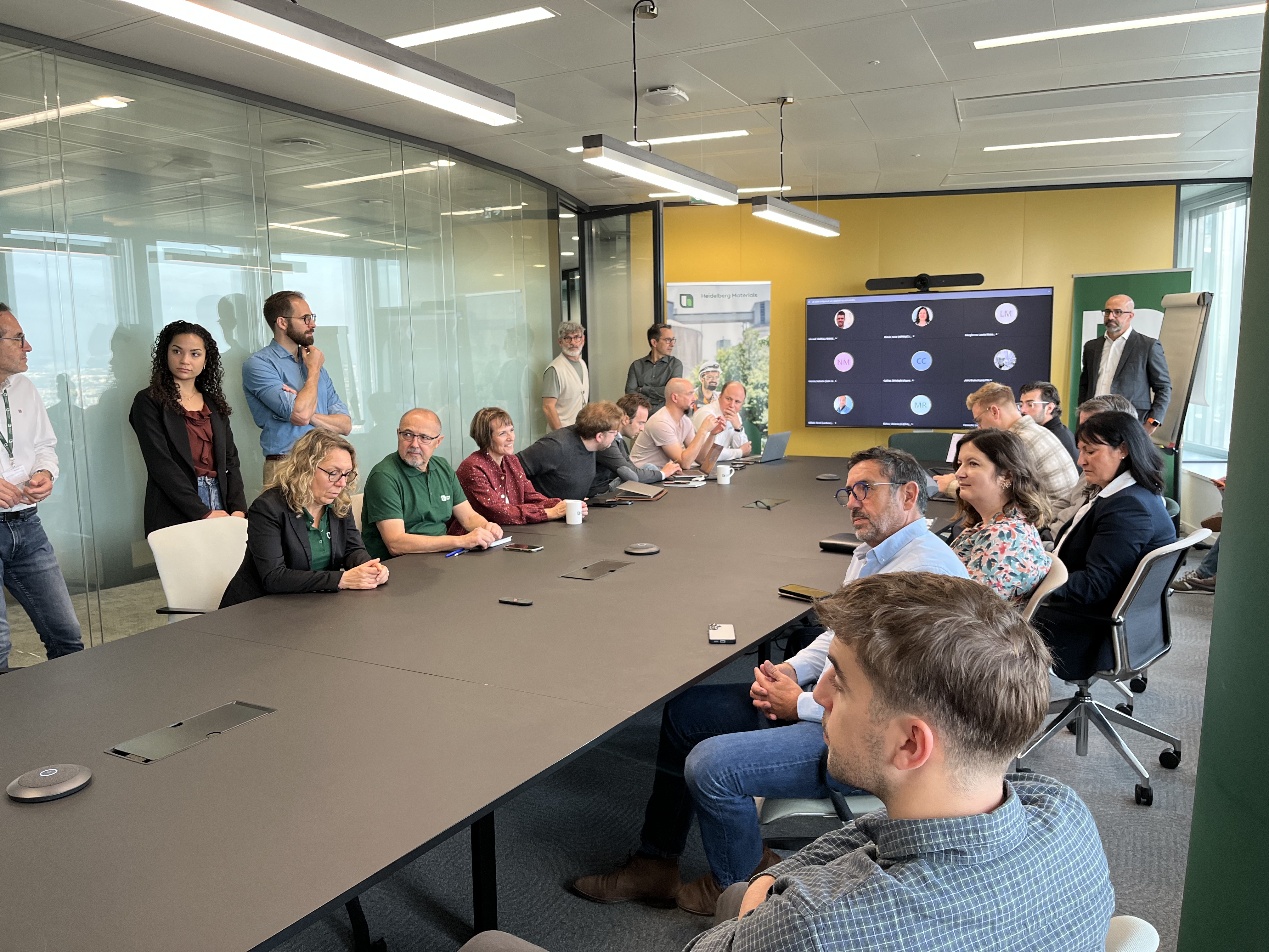 Des employés sont réunis autour d’une grande table de réunion dans une salle lumineuse avec un écran à droite affichant des avatars. Certains sont assis, d'autres debout à l'arrière, écoutant une présentation ou échangeant. Les murs sont vitrés à gauche et jaunes à droite.