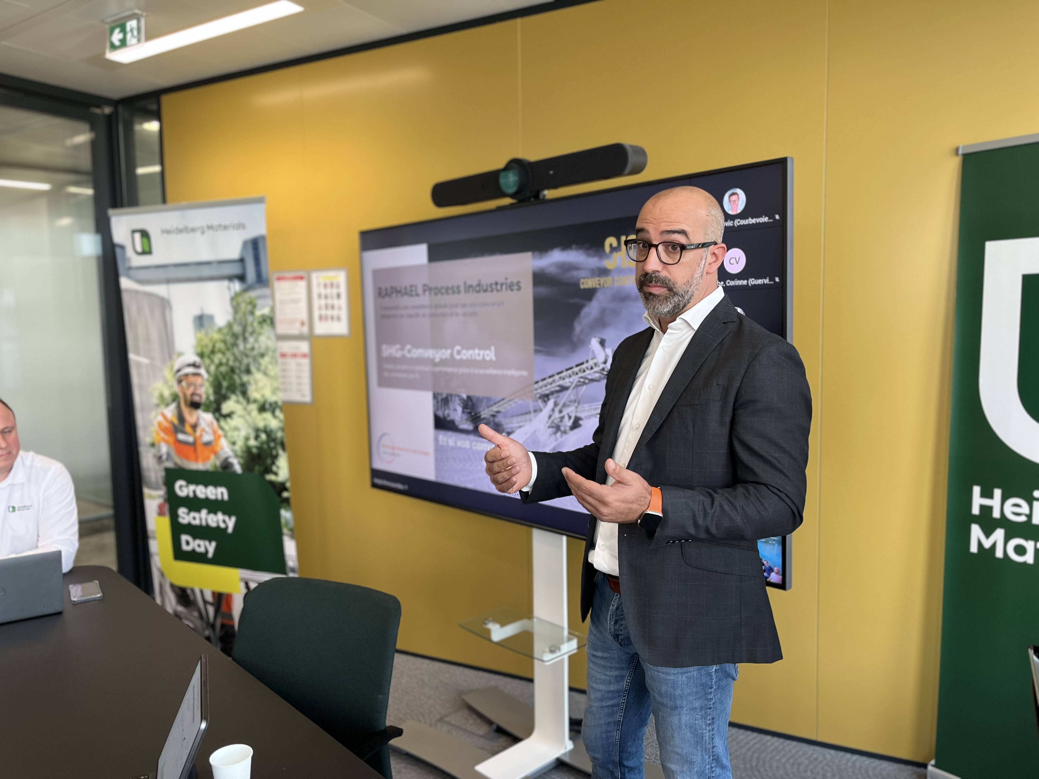 Un homme présente un contenu affiché sur un écran interactif dans une salle de réunion. L'écran montre des diapositives liées à un sujet industriel ou professionnel. À gauche, des affiches promotionnelles mentionnent "Green Safety Day" et le logo d'une entreprise est visible. La salle est lumineuse avec des équipements modernes.