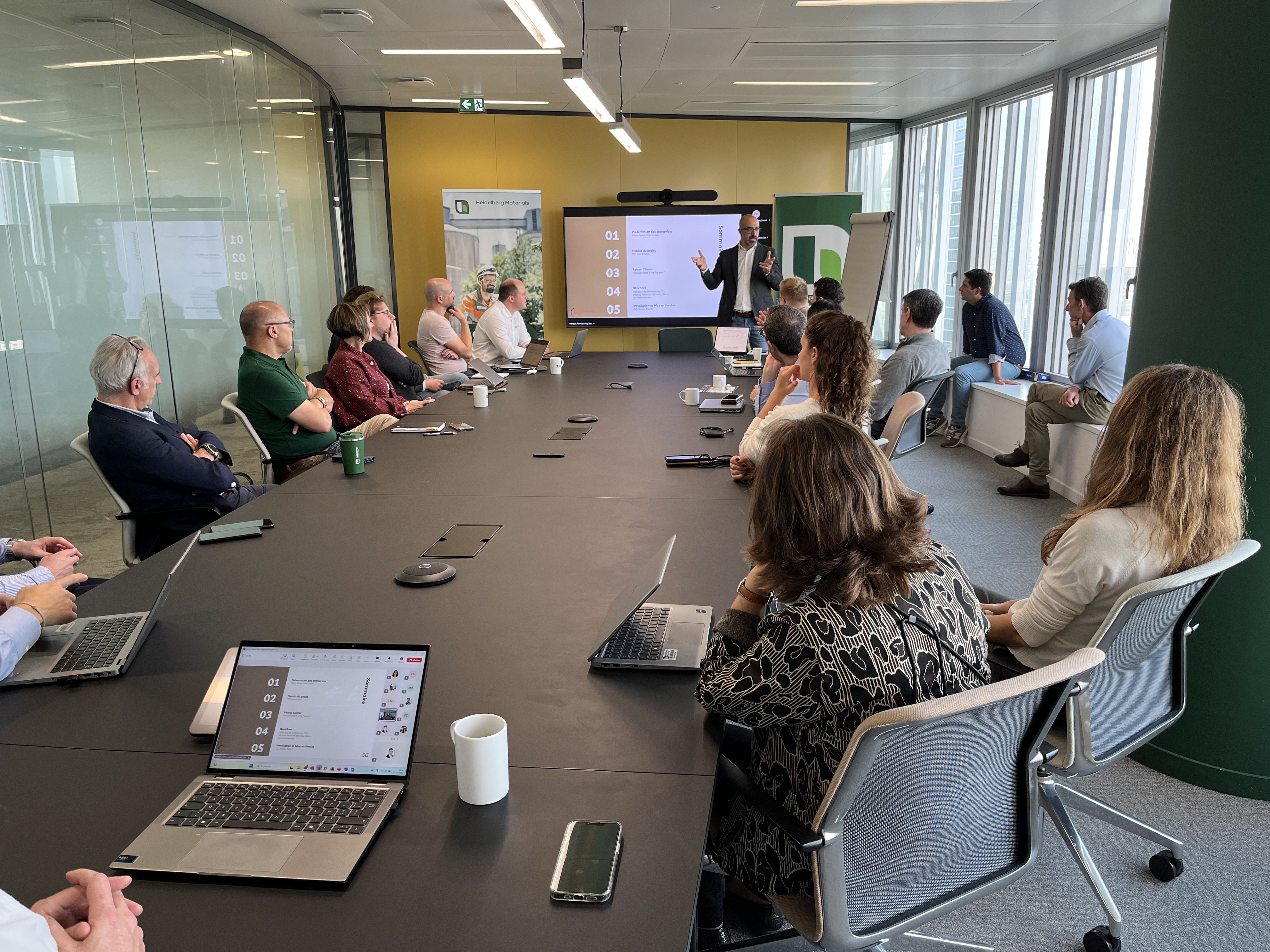Une réunion en salle de conférence avec des participants assis autour d'une longue table noire, certains utilisant des ordinateurs portables. Une personne debout à l'avant explique, pointant un écran affichant une présentation. Fenêtres vitrées sur la droite laissant entrer la lumière naturelle.