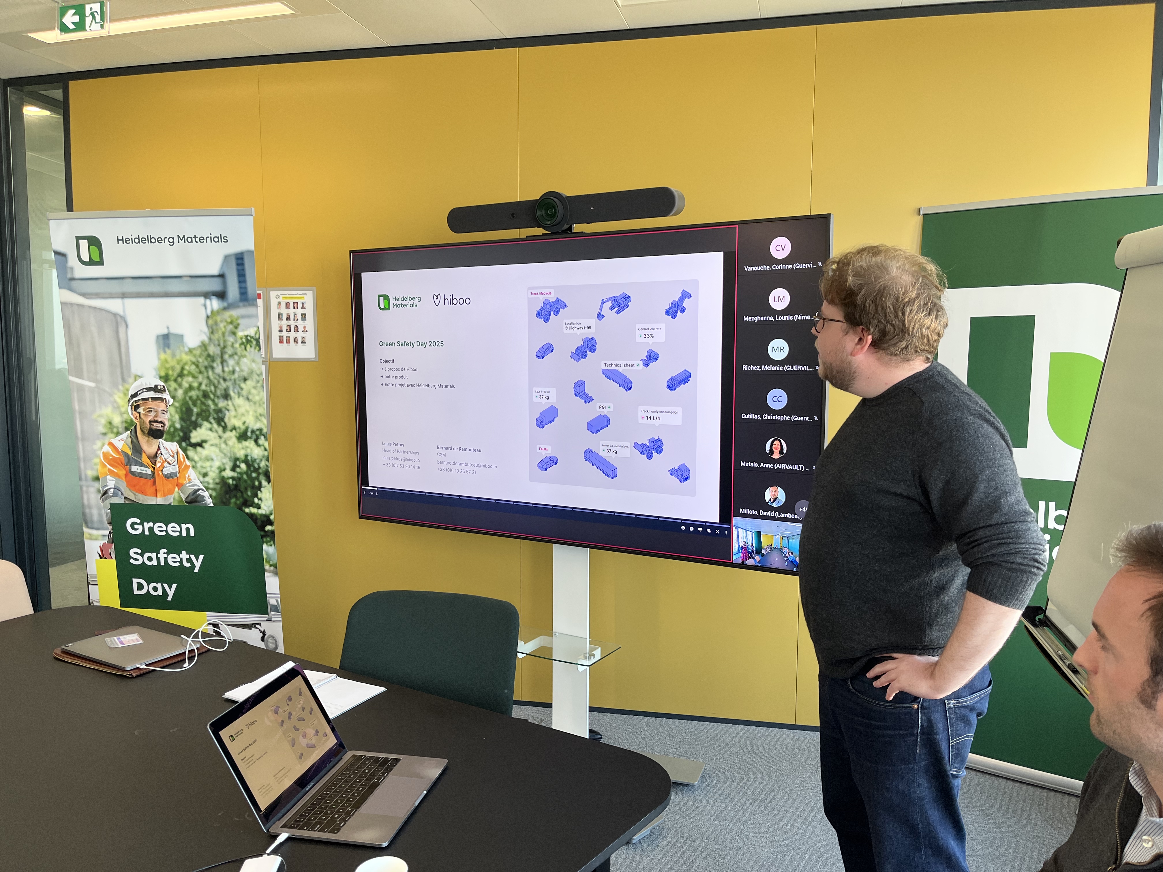 Un homme présente un écran interactif affichant un diagramme bleu et blanc, entouré par des participants assis. En arrière-plan, une affiche sur la sécurité environnementale et un tableau à feuilles mobiles sont visibles. La salle de réunion est éclairée par une lumière naturelle.