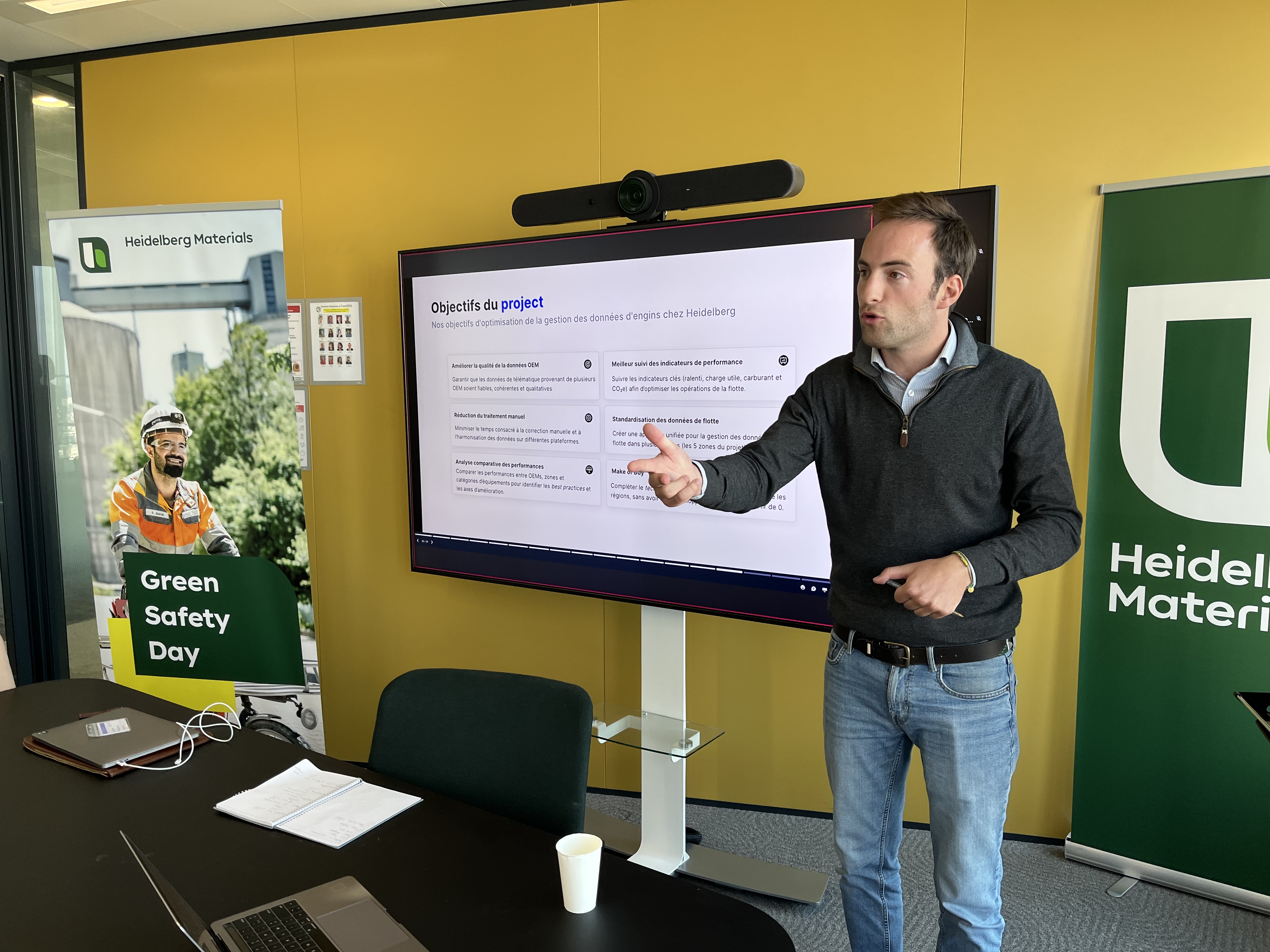 Un homme debout à droite, pointant vers un écran avec une présentation PowerPoint affichant des points clés. À gauche, un panneau sur la « Green Safety Day » et le logo de Heidelberg Materials. Fond de mur jaune, table de conférence avec des objets divers au premier plan.