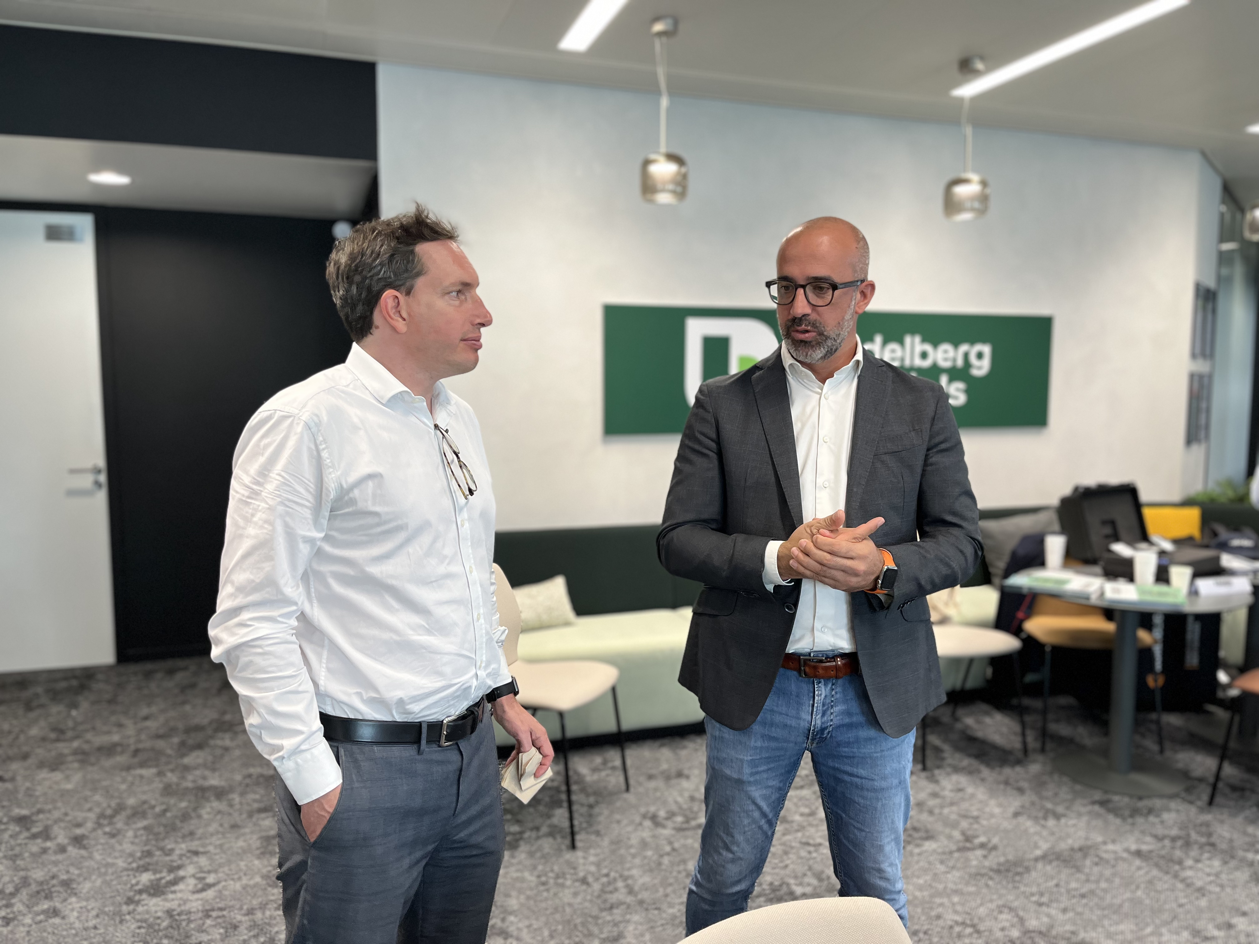 Deux hommes discutent dans un bureau moderne, avec un logo "Heidelberg" visible en arrière-plan sur un mur blanc. L'homme à gauche porte une chemise blanche et tient un objet, tandis que l'homme à droite, en veste grise et lunettes, gesticule pendant la conversation. Des chaises et une table sont visibles à droite.