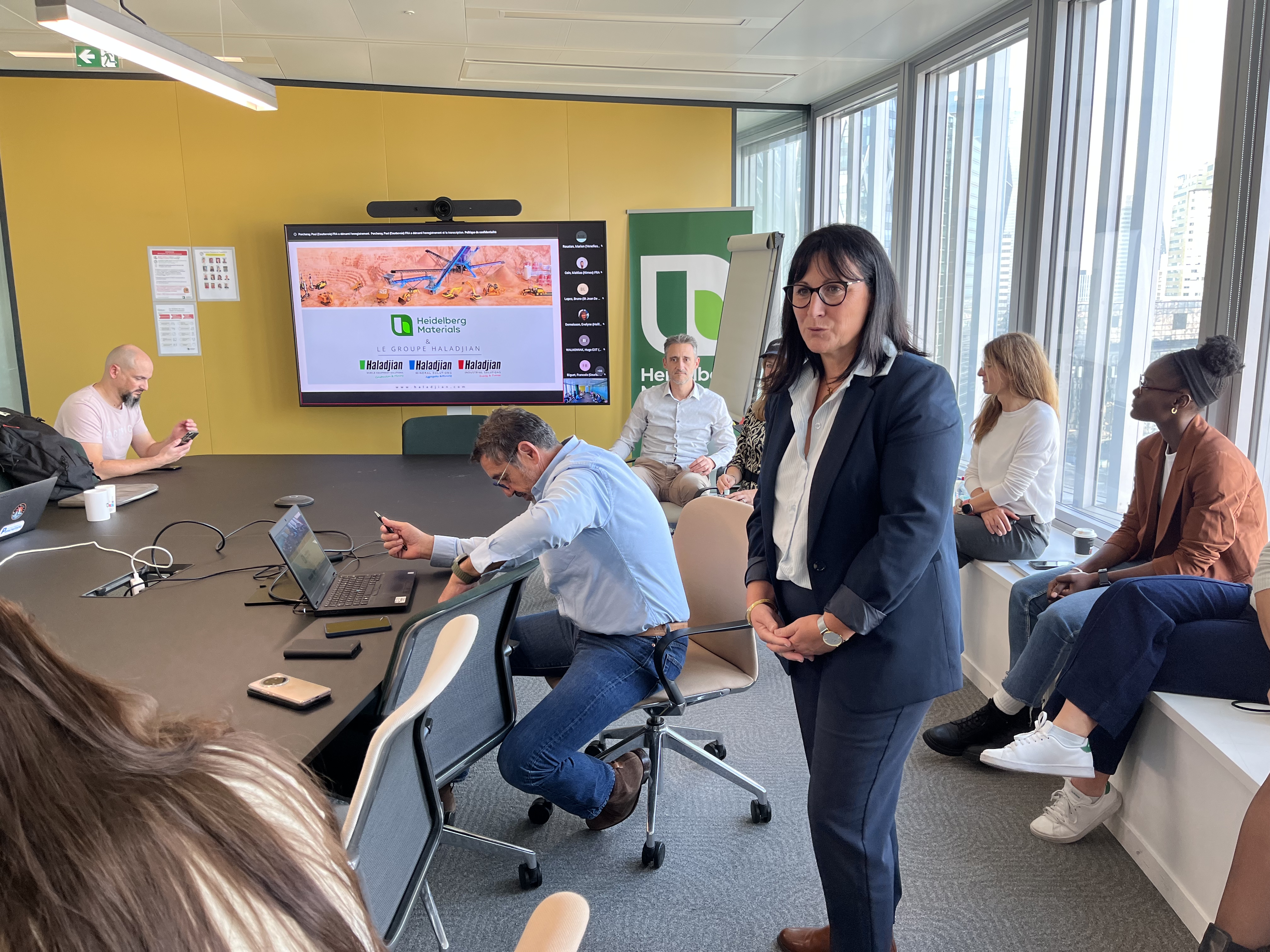 Femme en costume debout au premier plan dans une salle de réunion lumineuse, avec plusieurs participants assis, discutant ou utilisant des ordinateurs. En arrière-plan, un écran affiche une présentation sur "Green Finance Forum". Des fenêtres vitrées à droite laissent entrer la lumière naturelle.