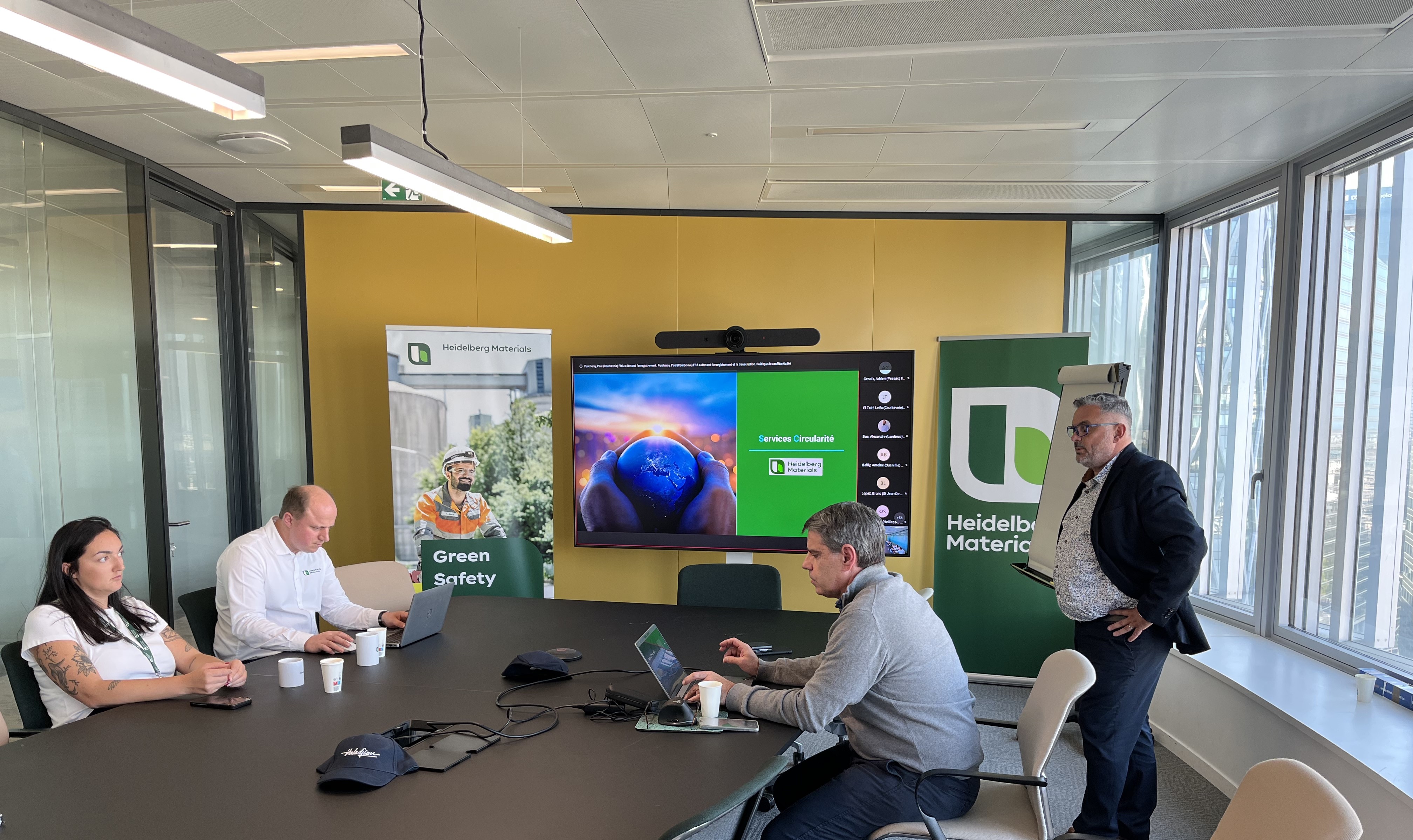 Quatre personnes sont réunies dans une salle de réunion avec une table noire au centre. À l'arrière-plan, un écran affiche une présentation colorée avec des boutons "Start" et "Join". Deux panneaux portant le logo de l'Université Catholique de Louvain (UCLouvain) sont visibles des deux côtés. De grandes fenêtres sur la droite laissent passer la lumière naturelle.